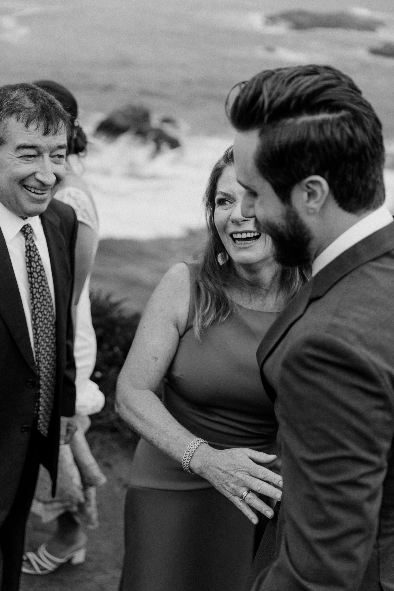Candid black and white photo of wedding guests sharing joyful laughter by the ocean.