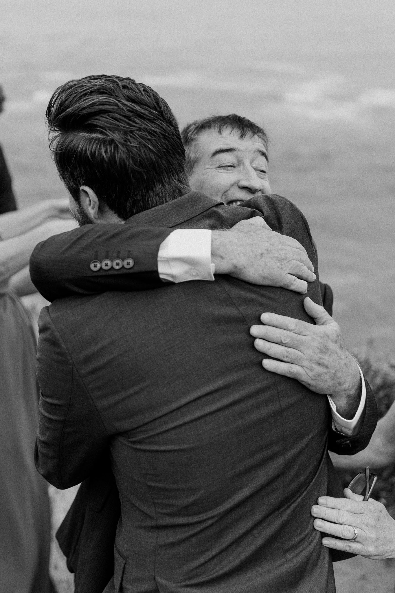Heartfelt black and white moment of two people embracing at coastal wedding ceremony.
