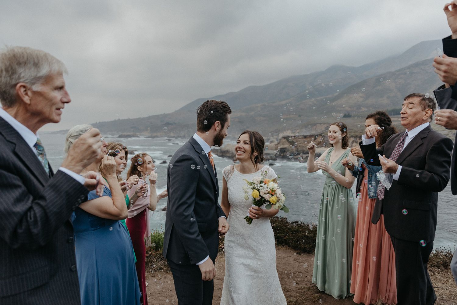 Wedding ceremony gathering on dramatic coastal cliff with guests watching the couple.