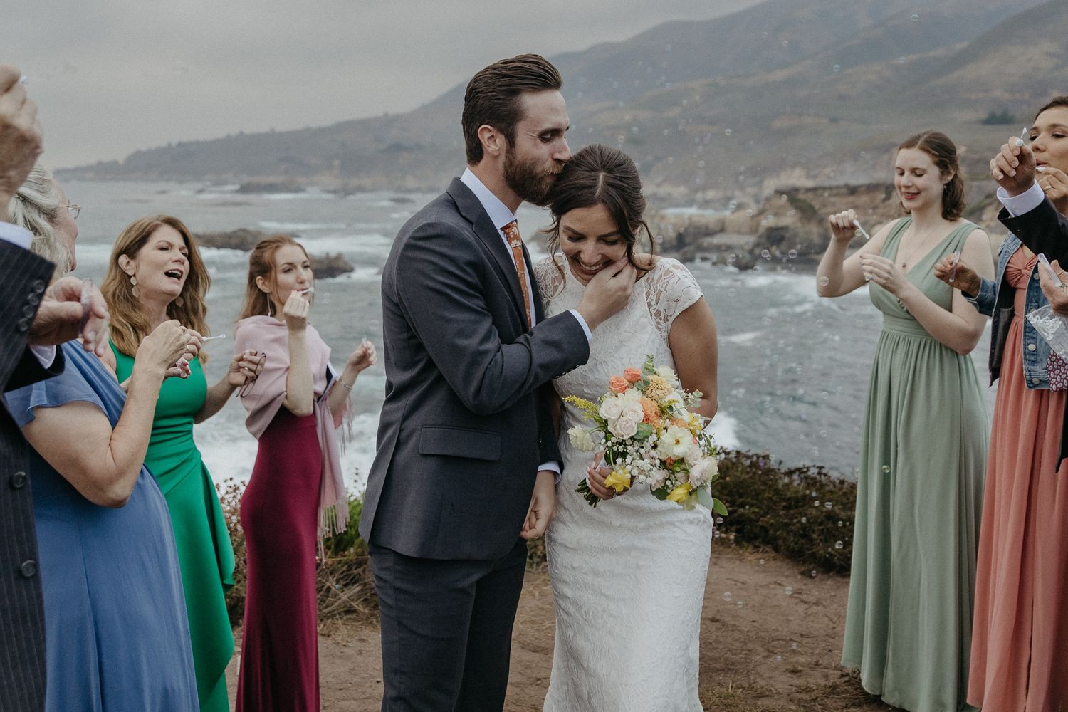 Wedding celebration on a scenic coastal cliff with guests in colorful dresses applauding the newlyweds.