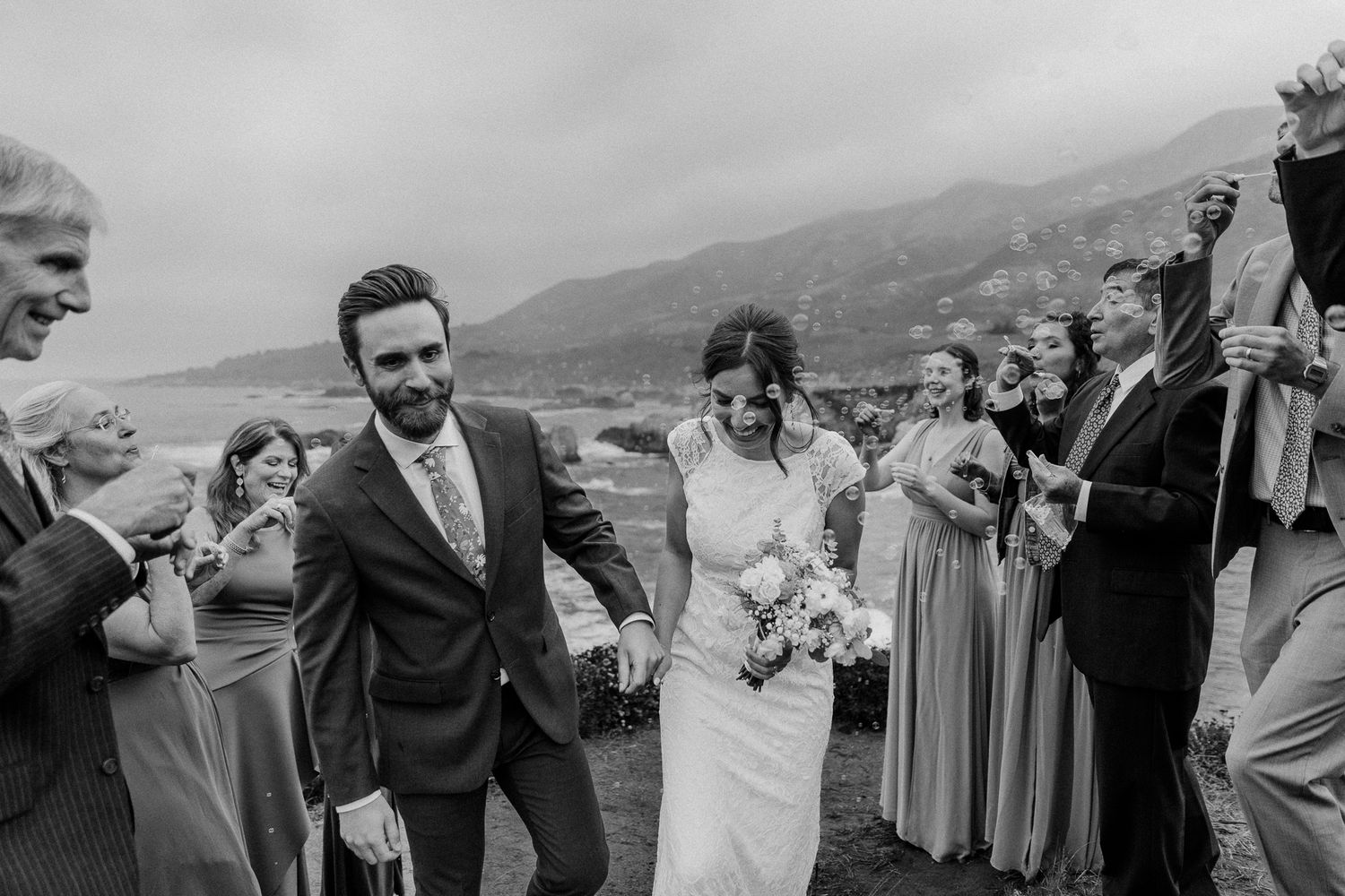 Wedding party celebrates with joyful laughter on a scenic mountainside in black and white.