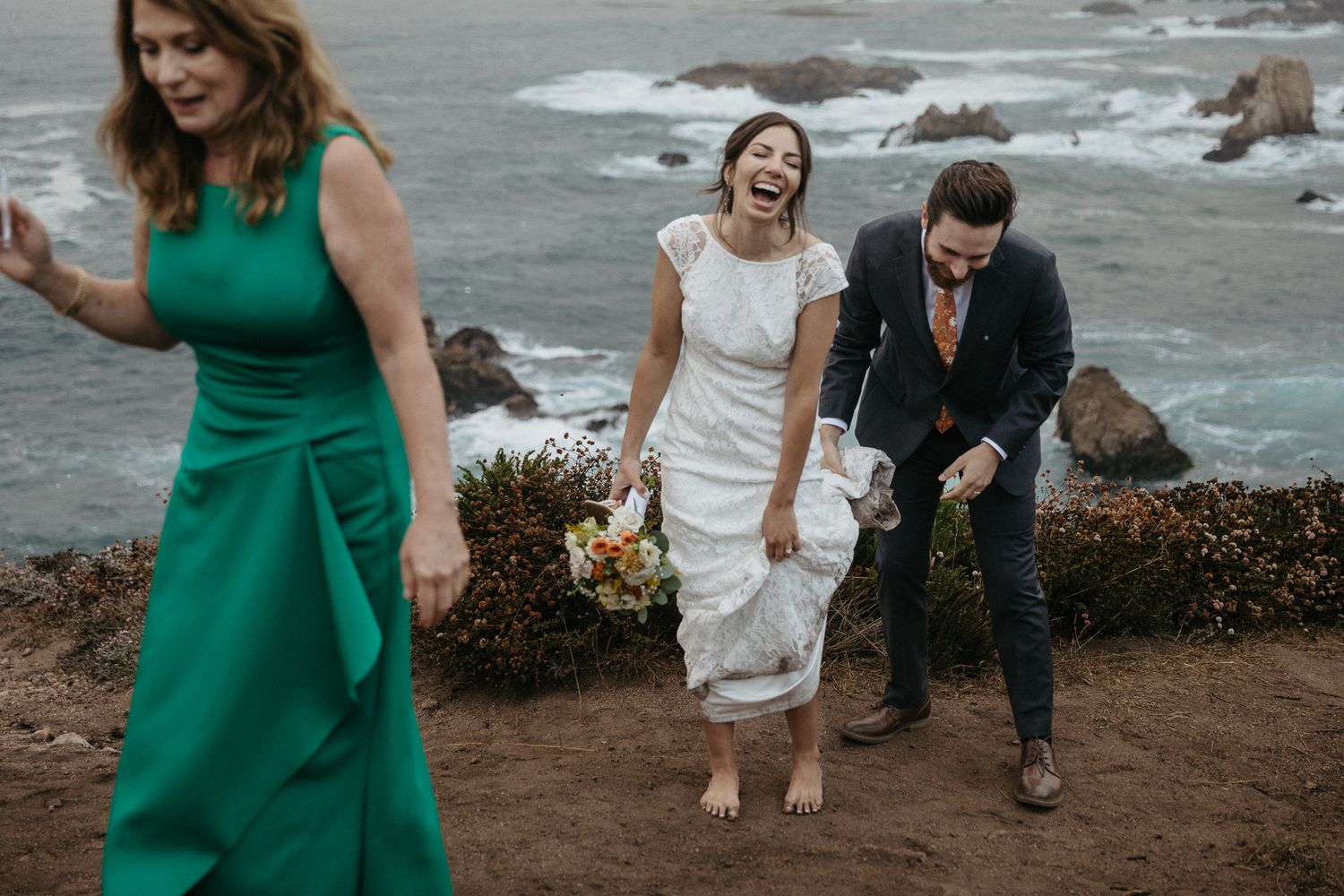 Wedding party laughs while walking along scenic coastal path with crashing waves below.