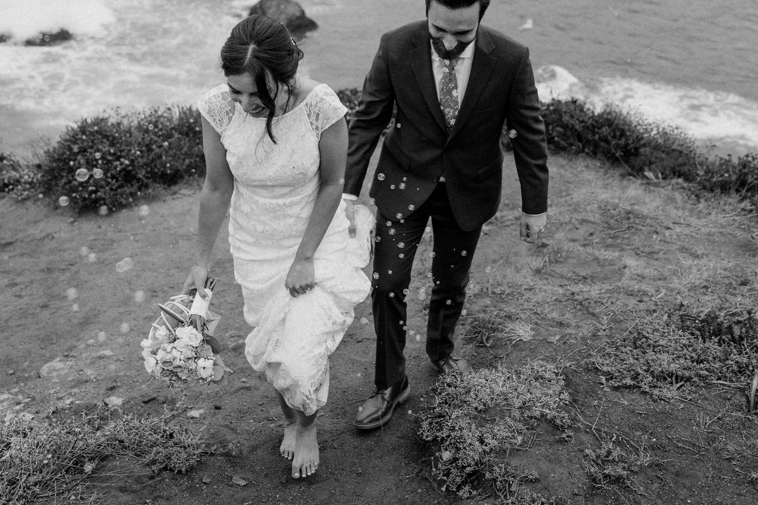 Newlyweds climb steep coastal path together in dramatic black and white photography.