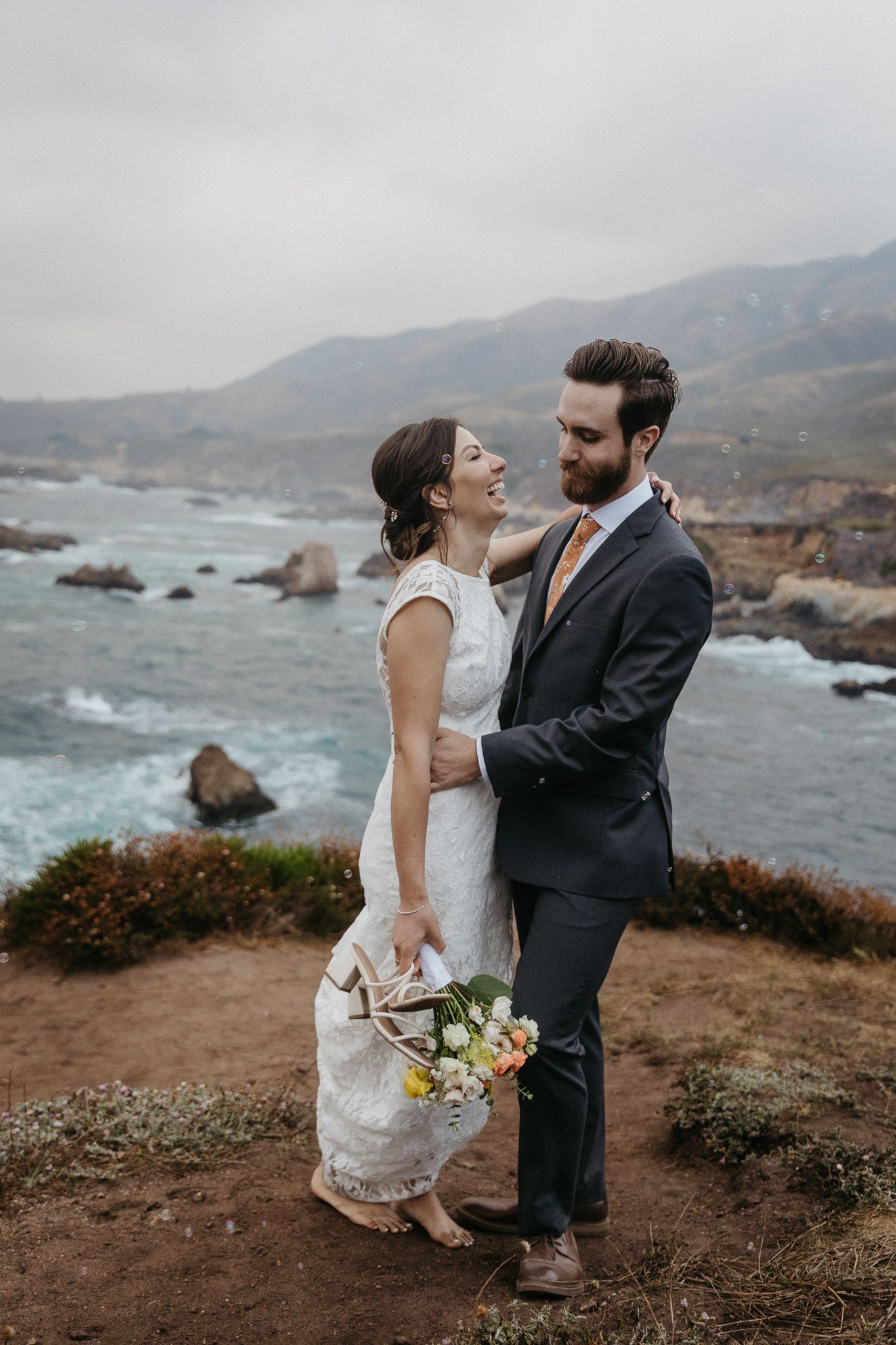 Romantic coastal wedding portrait with dramatic ocean and mountain backdrop.