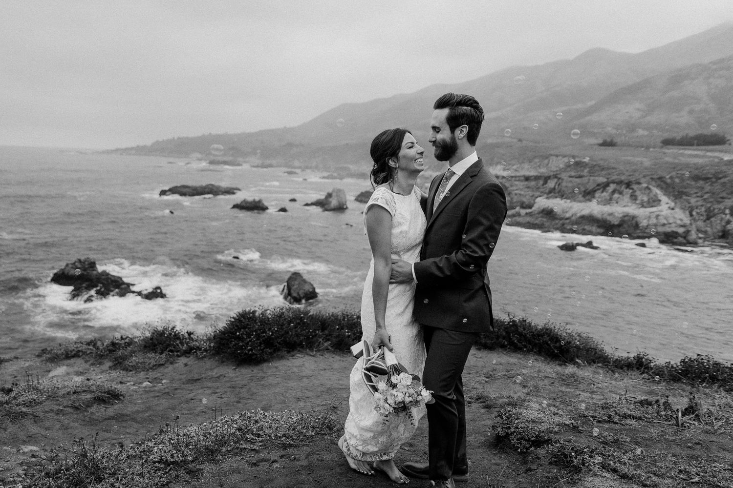 Black and white wedding portrait captures tender moment on scenic coastal overlook.