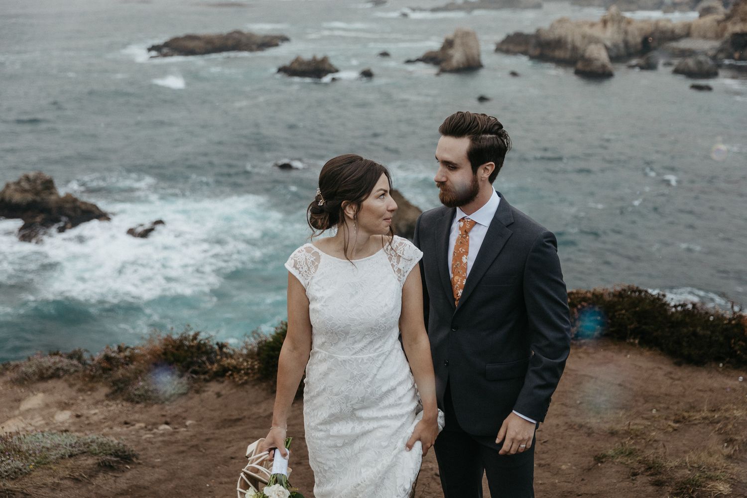 Elegant wedding couple shares intimate moment on cliff overlooking rocky coastline.