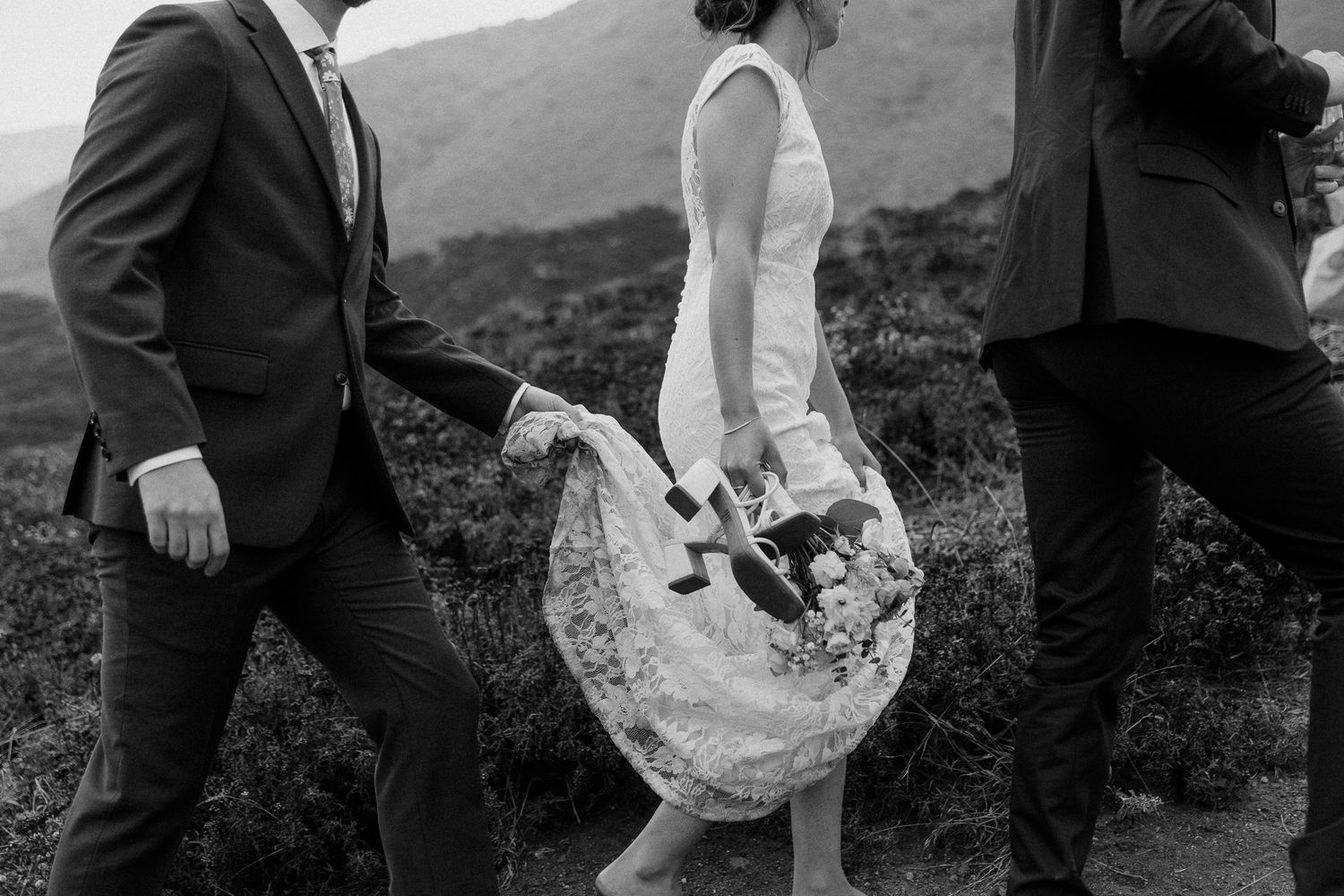 Wedding party helps lift bride's dress while walking on rugged coastal path.