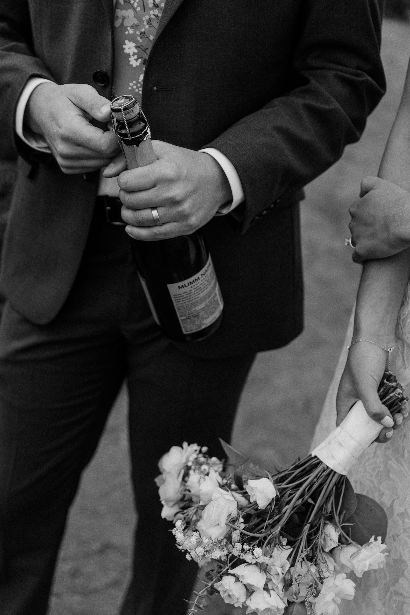Black and white photo of hands opening champagne bottle with flowers held in the foreground.