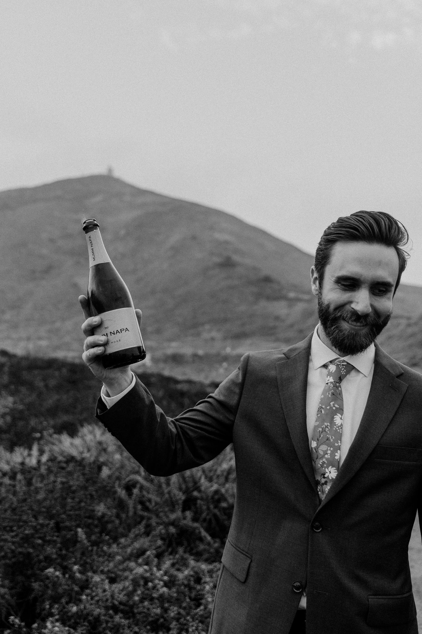 Man in suit holds up champagne bottle against mountainous backdrop.