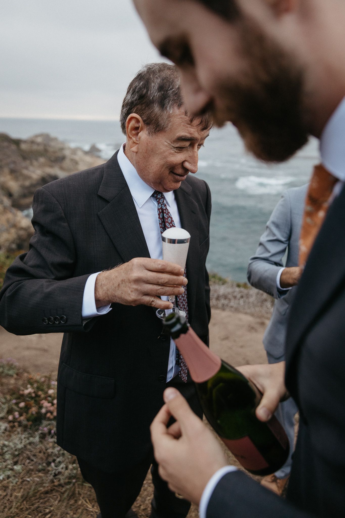 Wedding guests sharing drinks and laughter with ocean view backdrop.