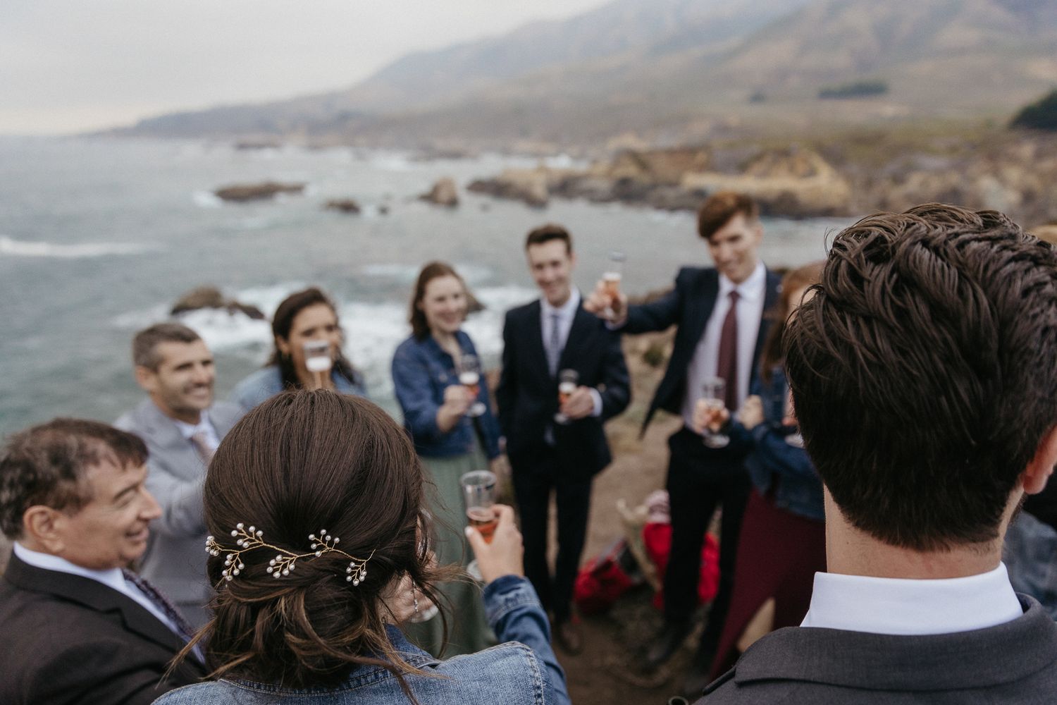 Wedding party gathered on coastal cliff for celebratory drinks.