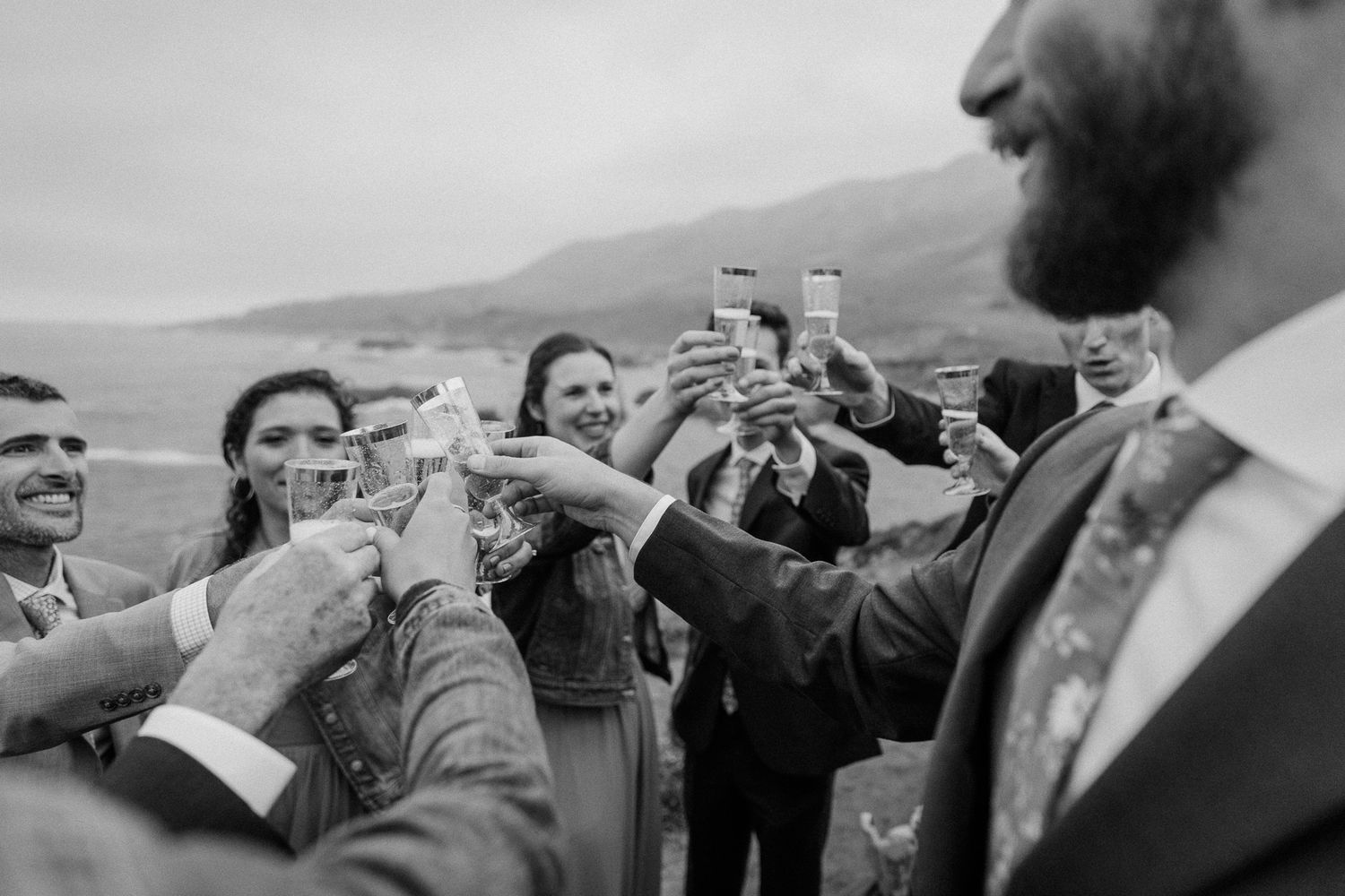 Wedding guests raise champagne glasses in a celebratory toast against a mountainous backdrop.