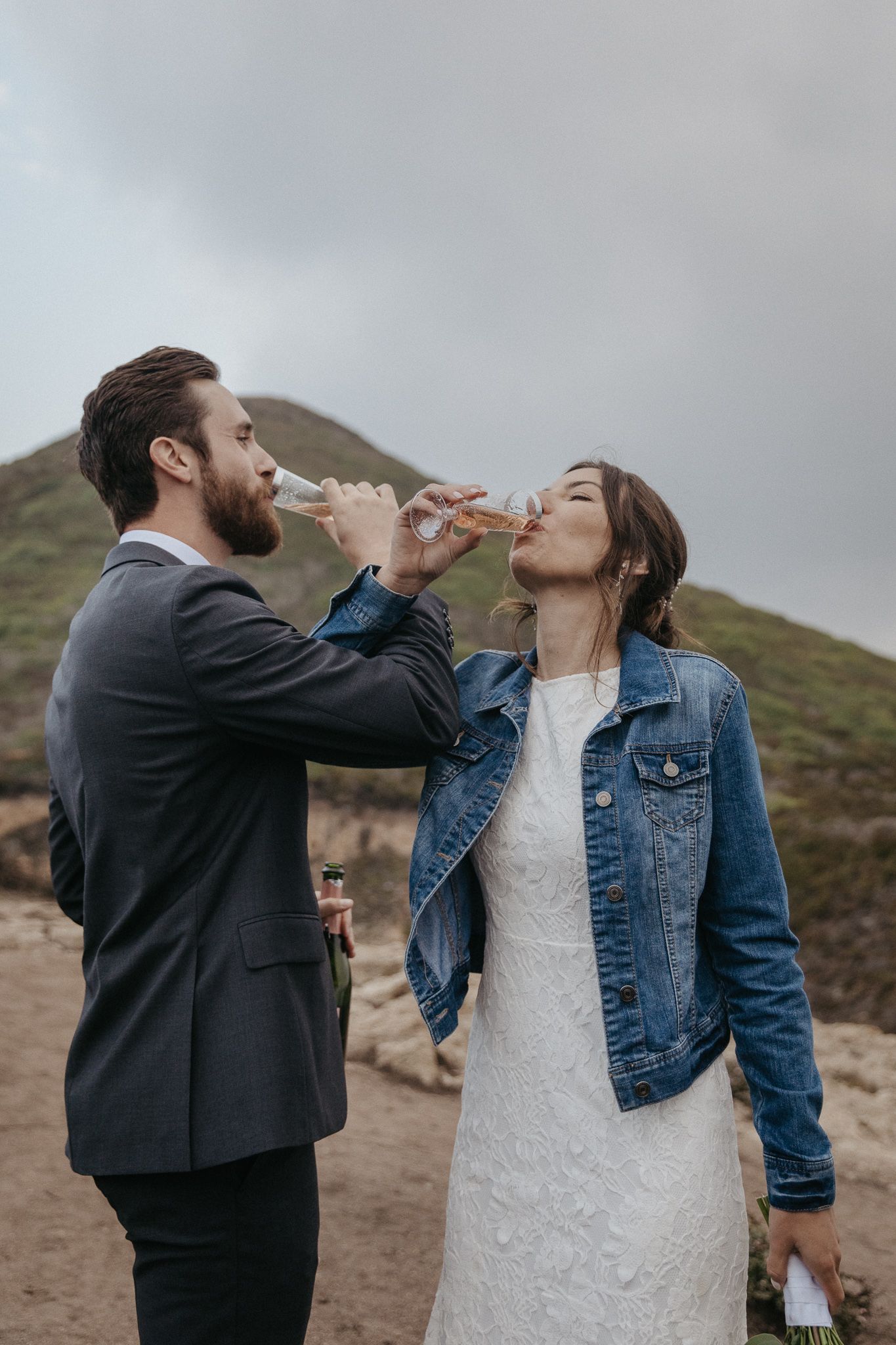 A couple in wedding attire share a playful champagne toast on a hilltop.