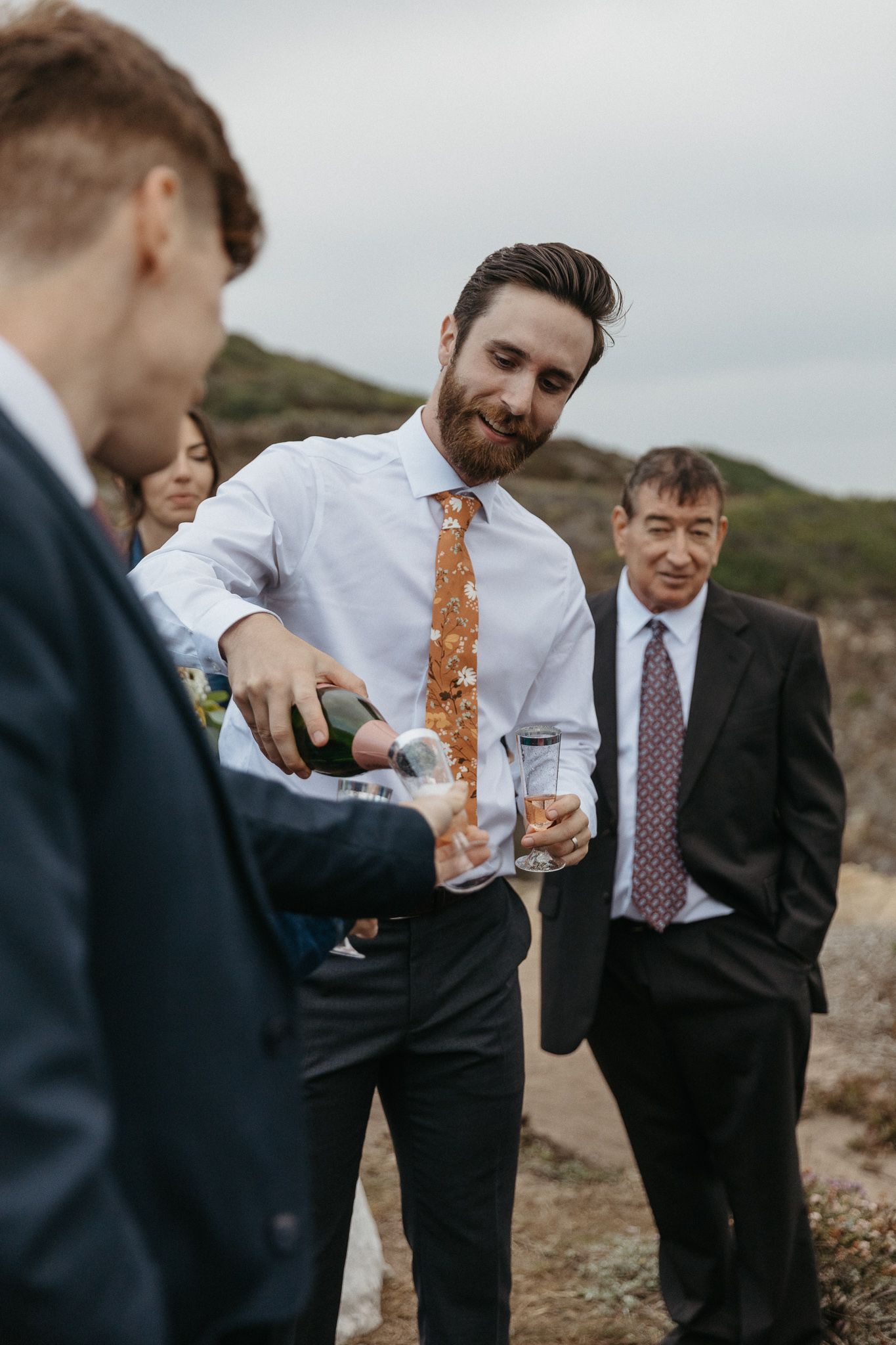 A person in formal attire pours drinks for guests at an outdoor celebration.