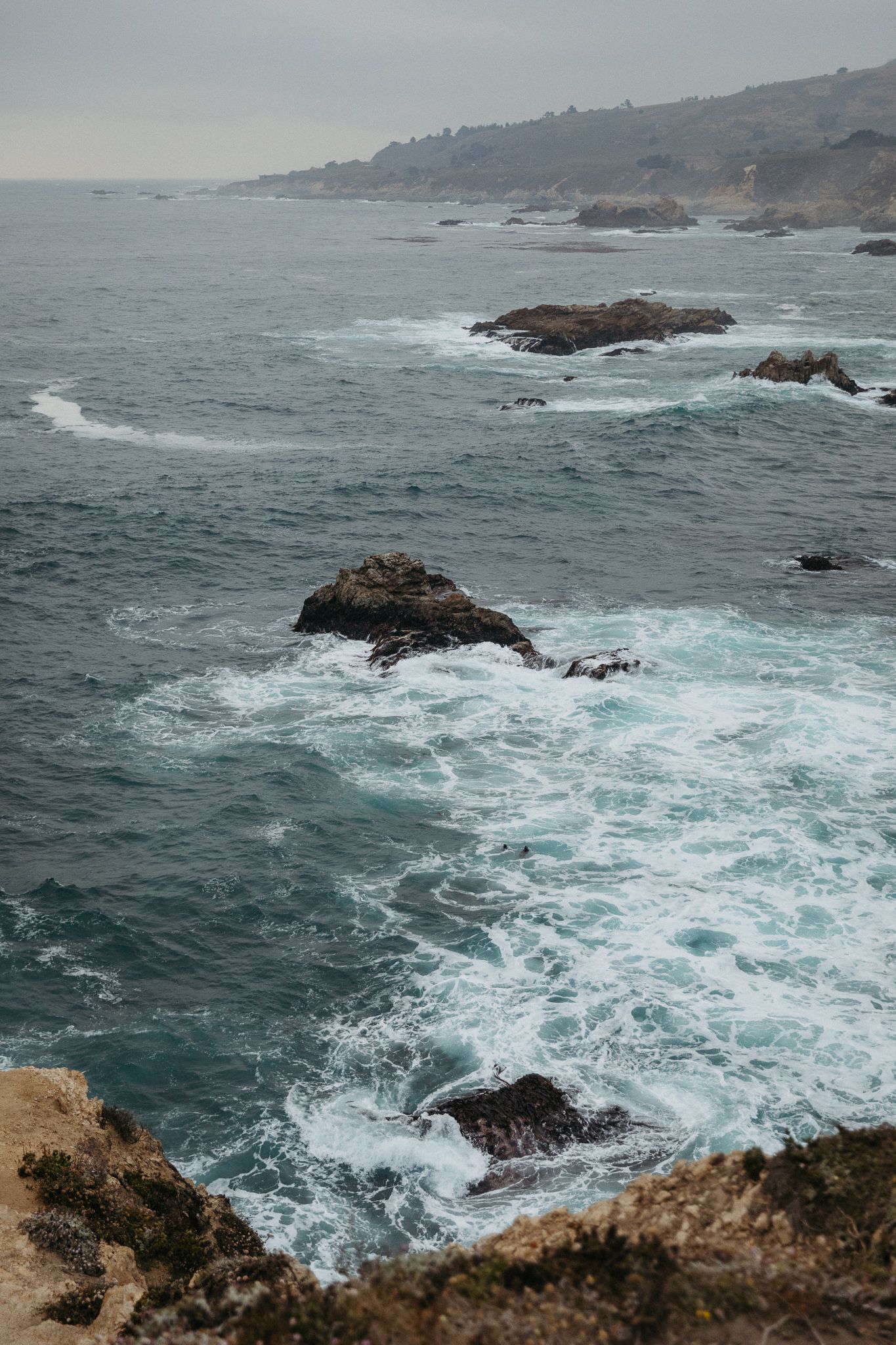 Rugged California coastline with waves crashing against rocky outcrops under cloudy skies.