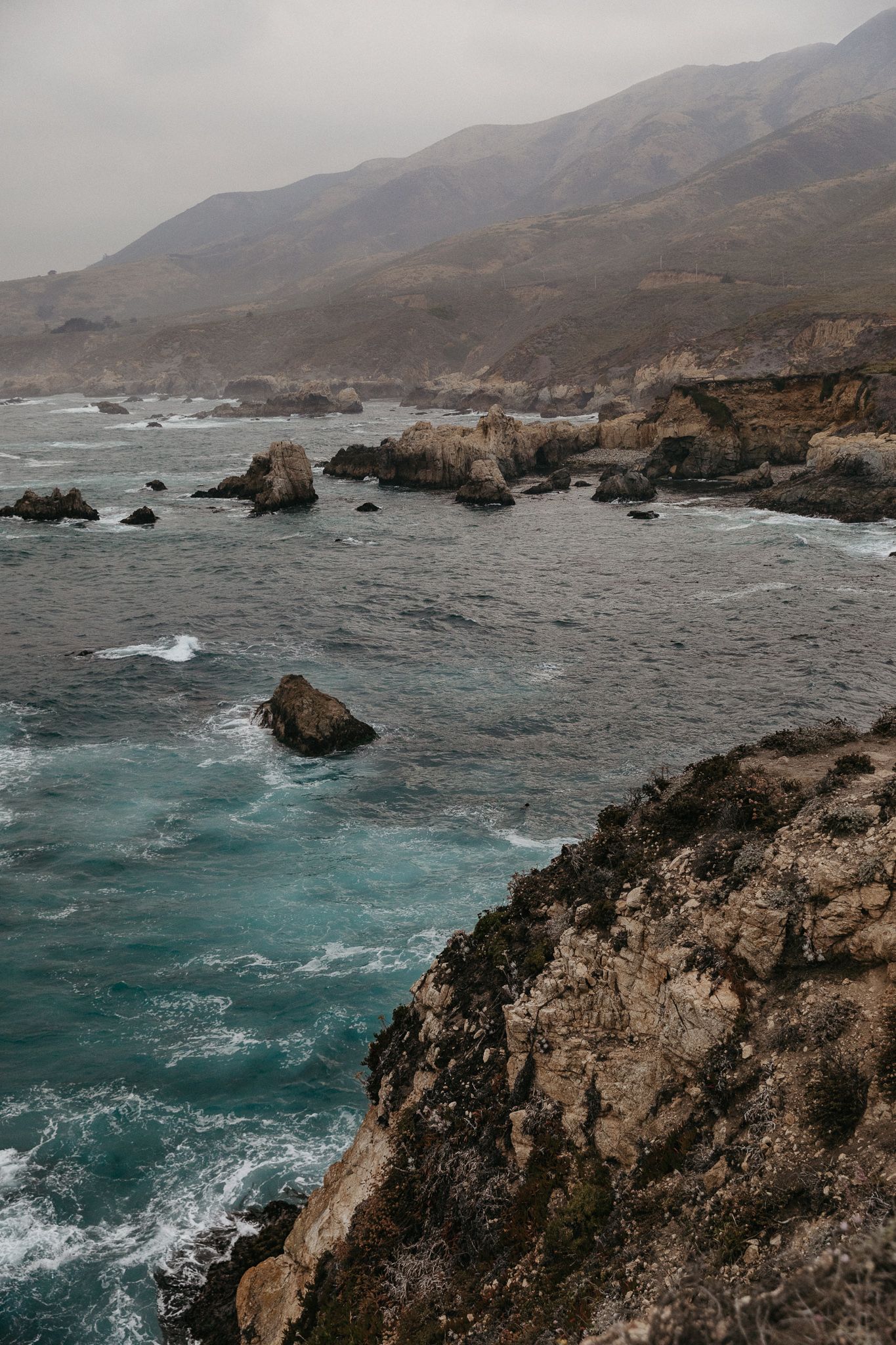 Dramatic view of Big Sur coastline with waves crashing against rocky cliffs under moody skies.