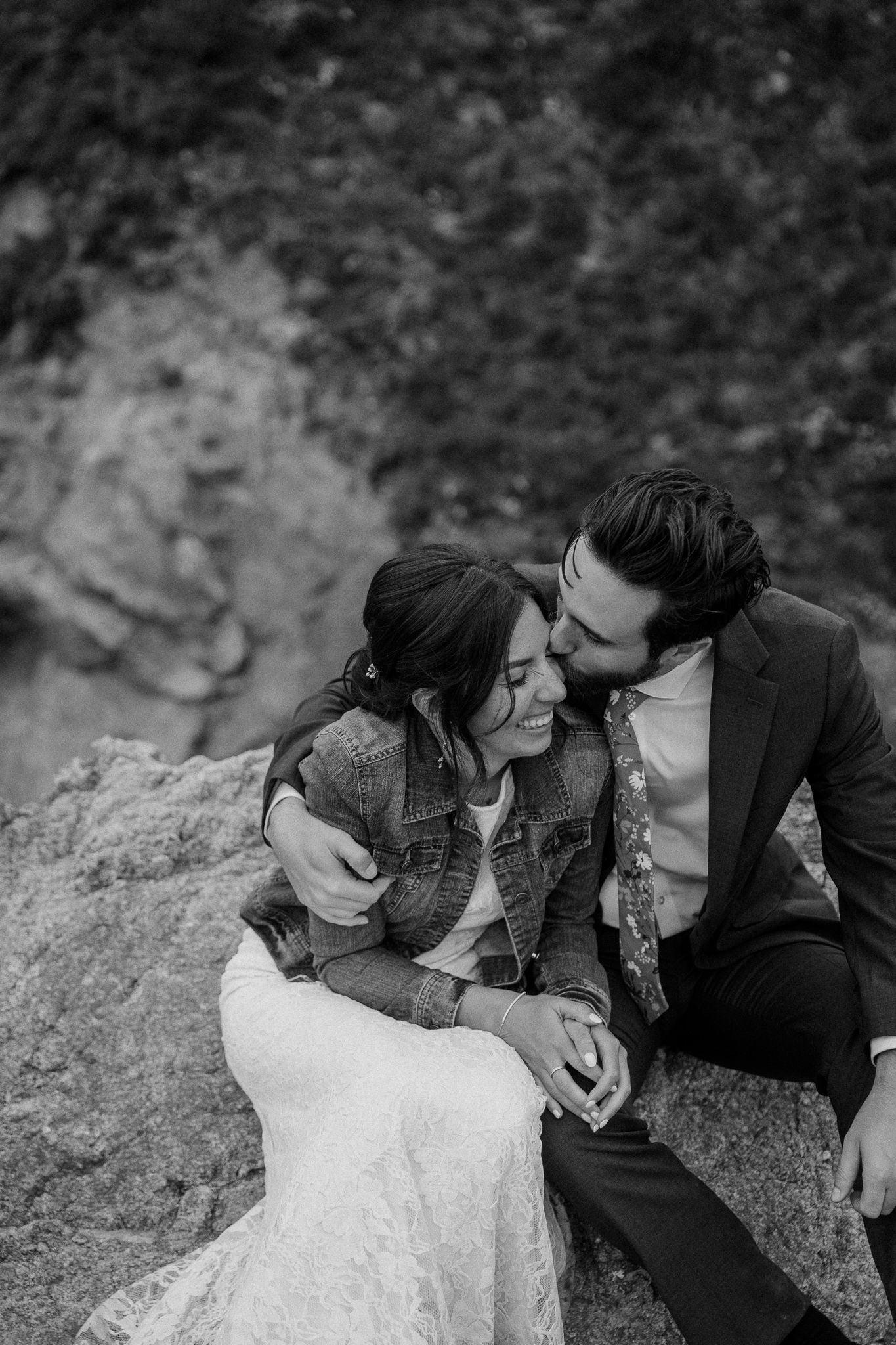 A romantic couple in wedding attire embrace on a rocky cliff wearing denim and formal wear.