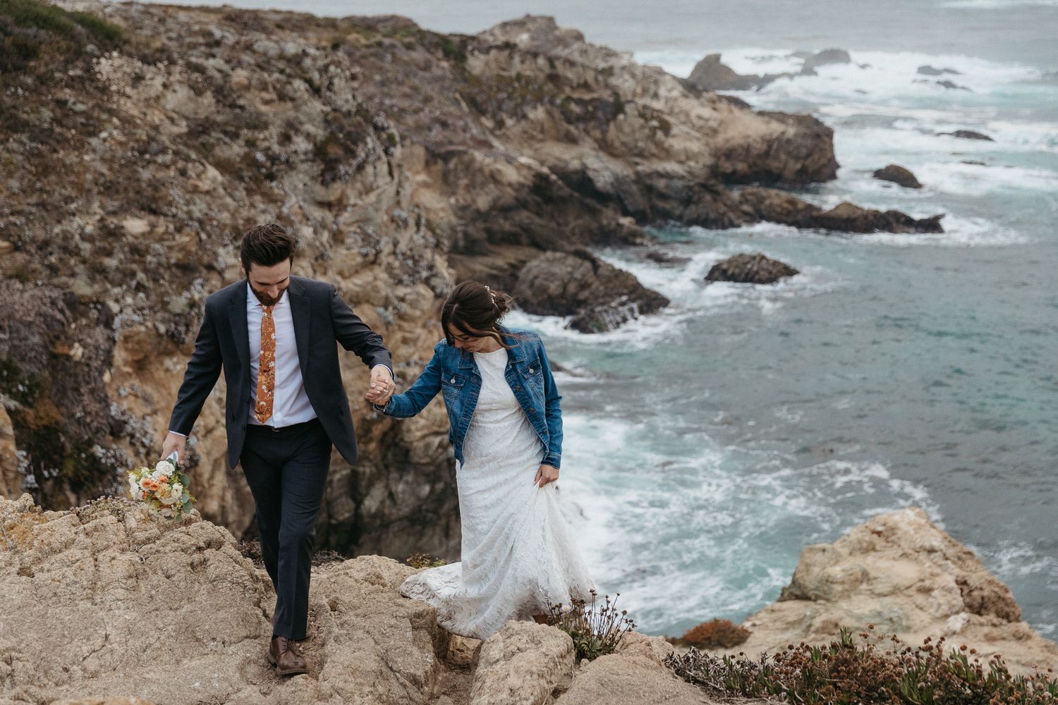 A couple walks along a dramatic coastal cliff path in wedding attire.