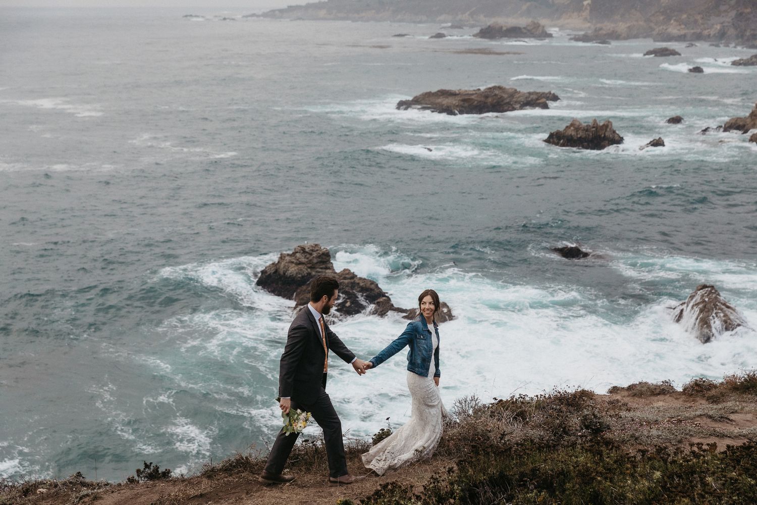 A couple holds hands while exploring a rugged oceanside cliff during their wedding.
