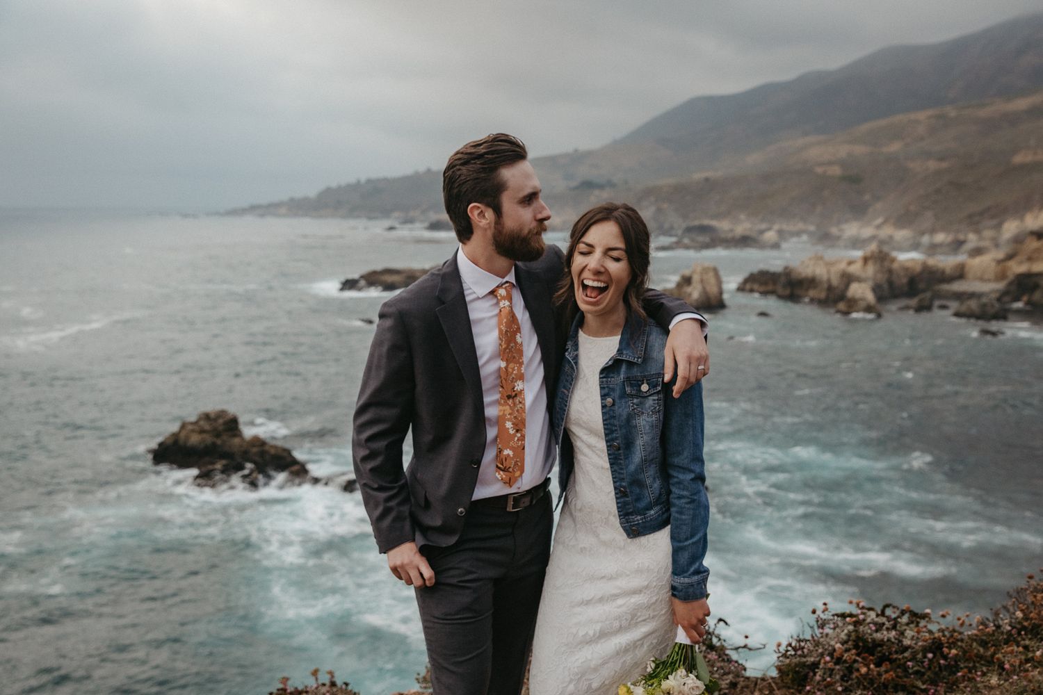A couple laughs together on a clifftop overlooking the dramatic Big Sur coastline.