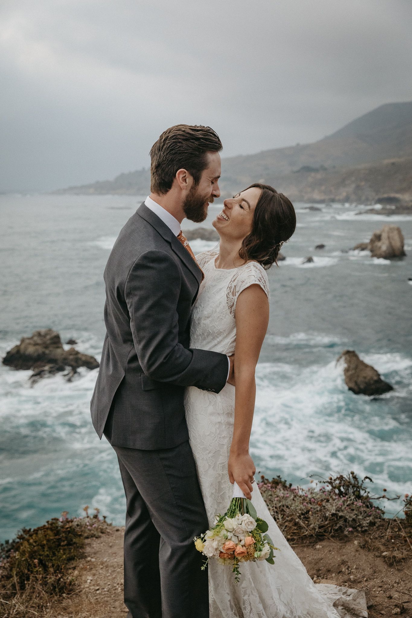 A romantic wedding portrait captured on a dramatic coastal cliff with crashing waves below.