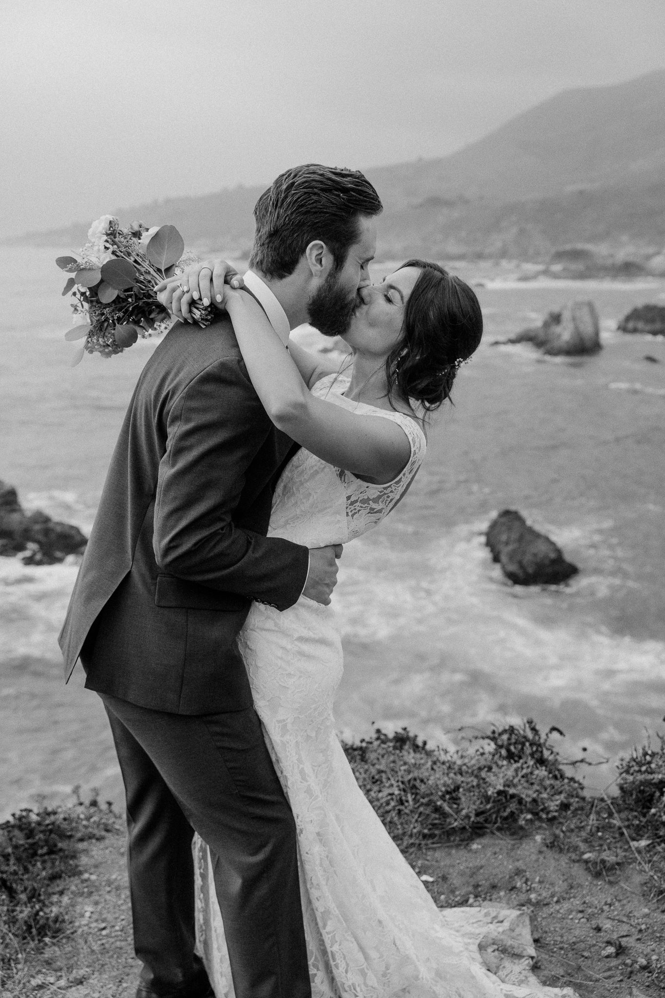 A romantic dip kiss between newlyweds on a cliff overlooking the ocean.