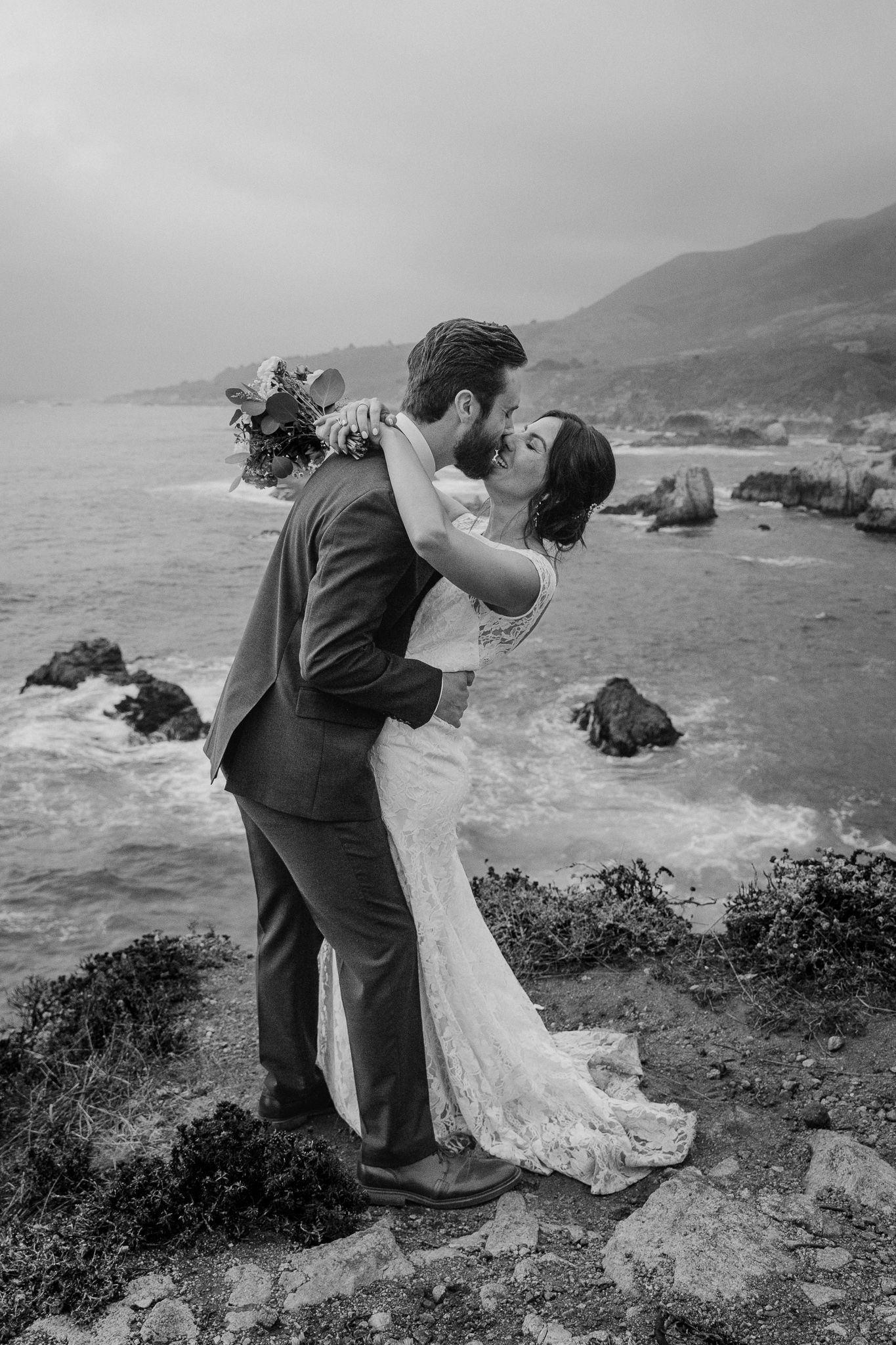 A dramatic black and white wedding portrait shows a couple's intimate moment on a coastal cliff.
