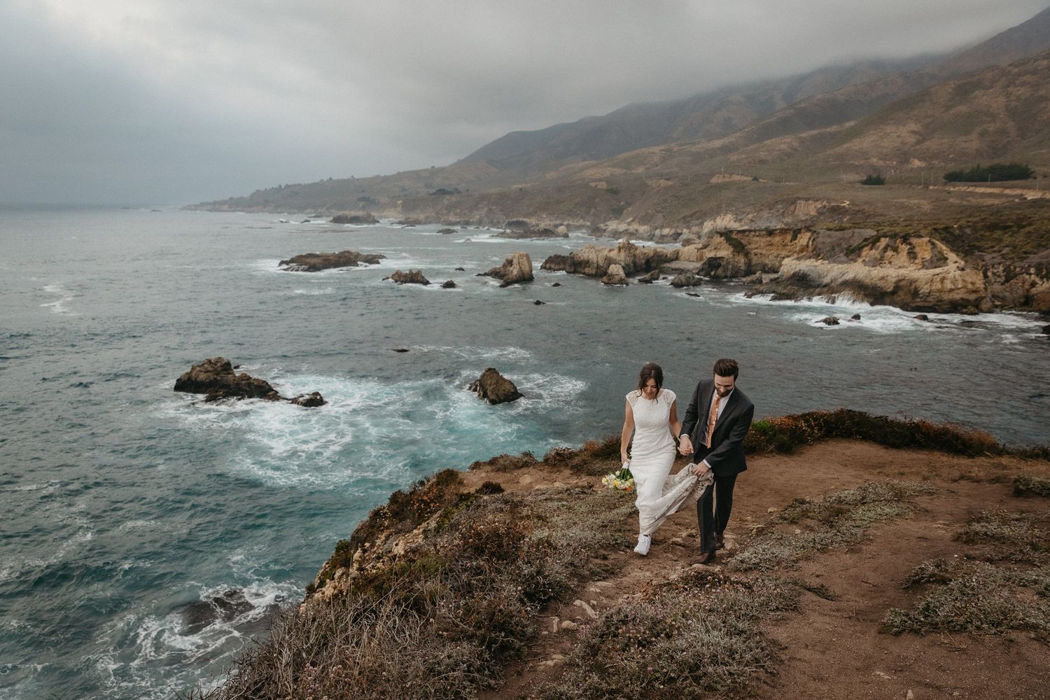 A wedding couple stands on a dramatic overlook with sweeping views of the Big Sur coastline.