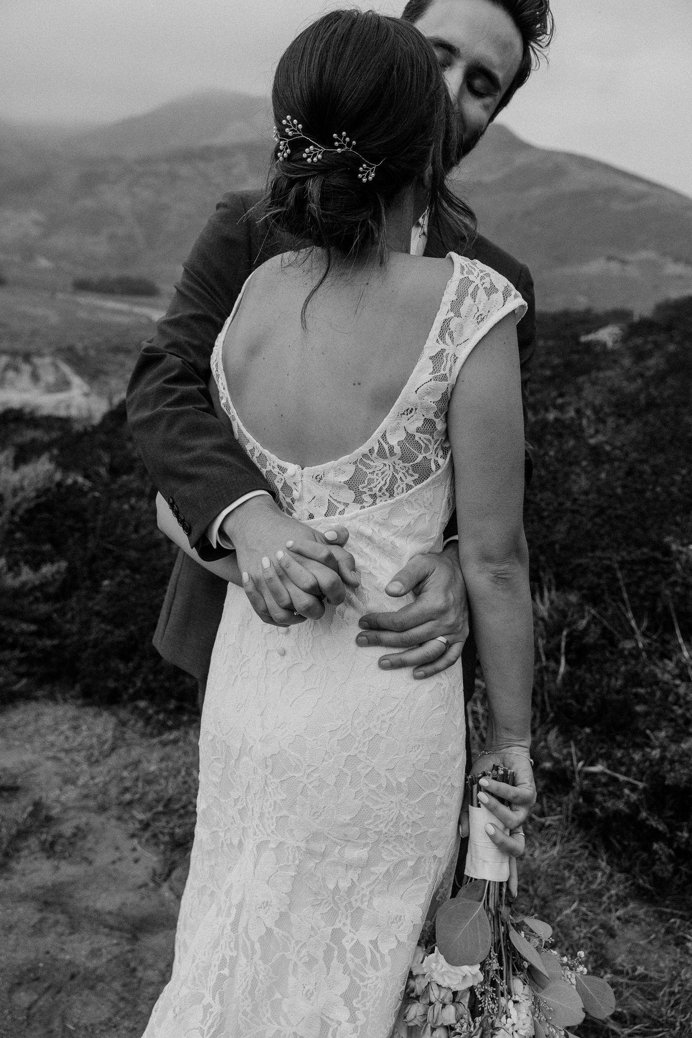 An intimate black and white portrait shows a couple's tender embrace against coastal mountains.
