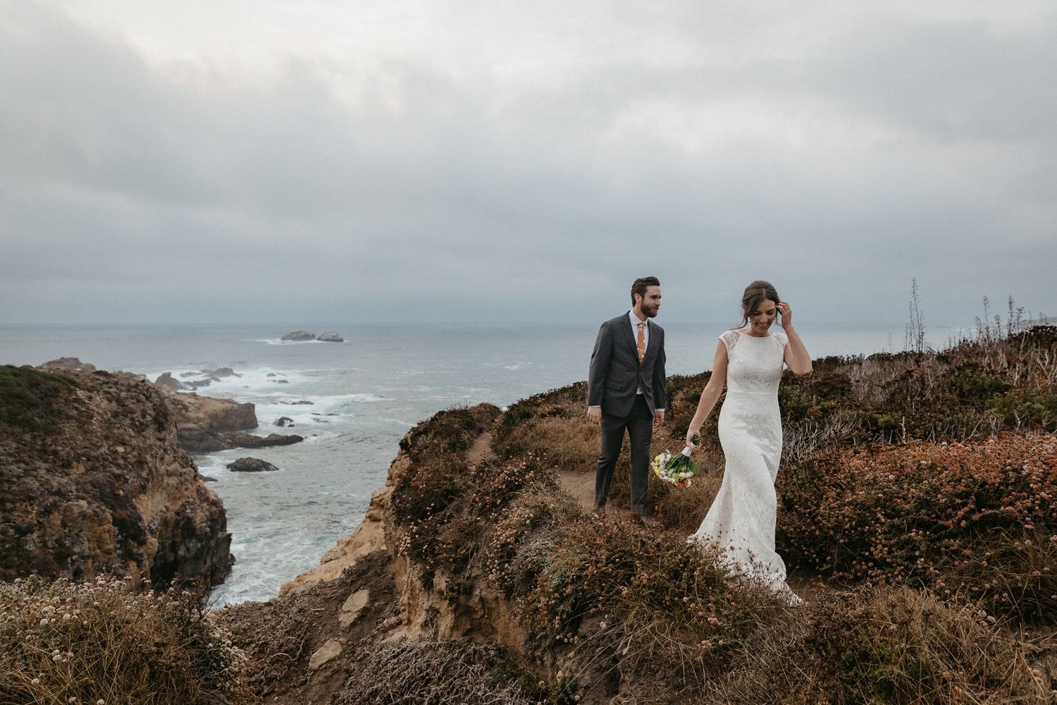A couple in wedding attire stands atop dramatic coastal cliffs overlooking a misty ocean landscape.