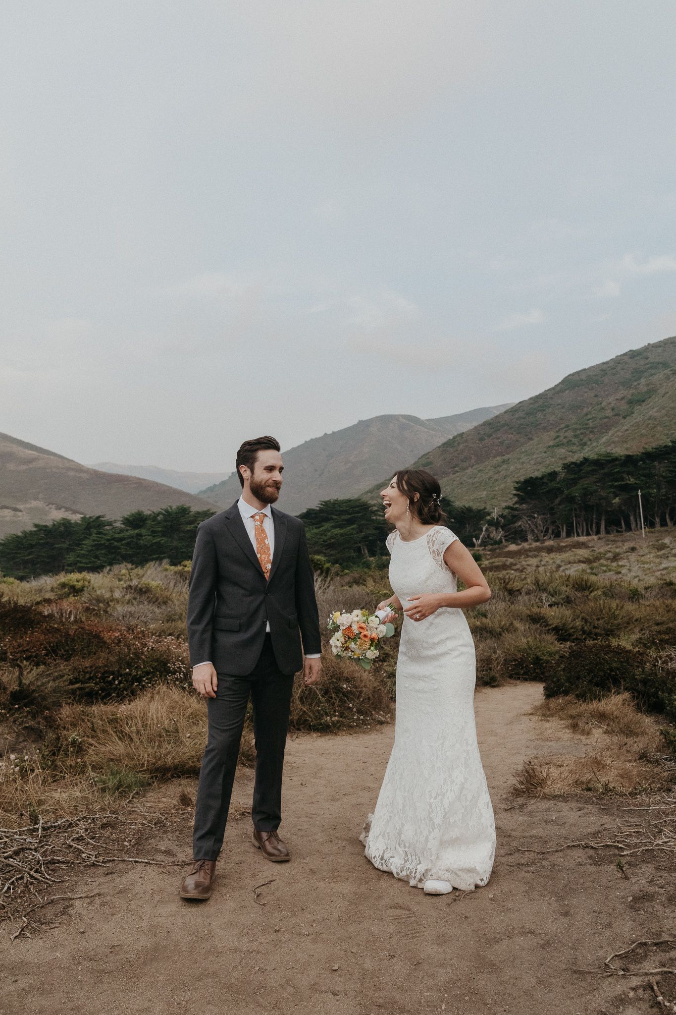 A couple shares a candid moment on a dirt path with rolling hills and mountains in the background.