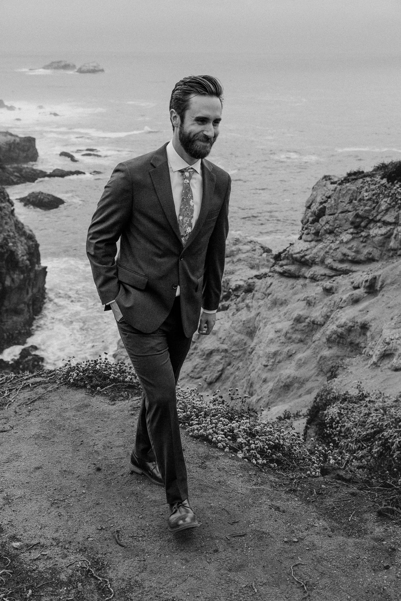 A suited figure walks along a dramatic coastal cliff path with crashing waves below.