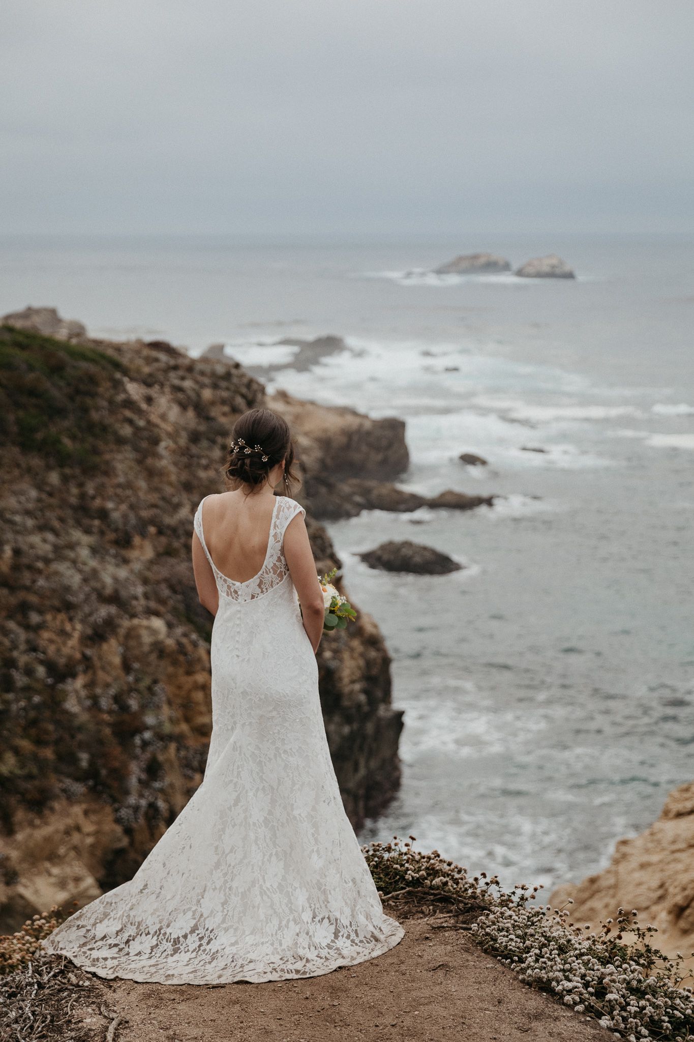 A bride in a lace gown stands at the edge of a coastal cliff, overlooking the rocky shoreline.