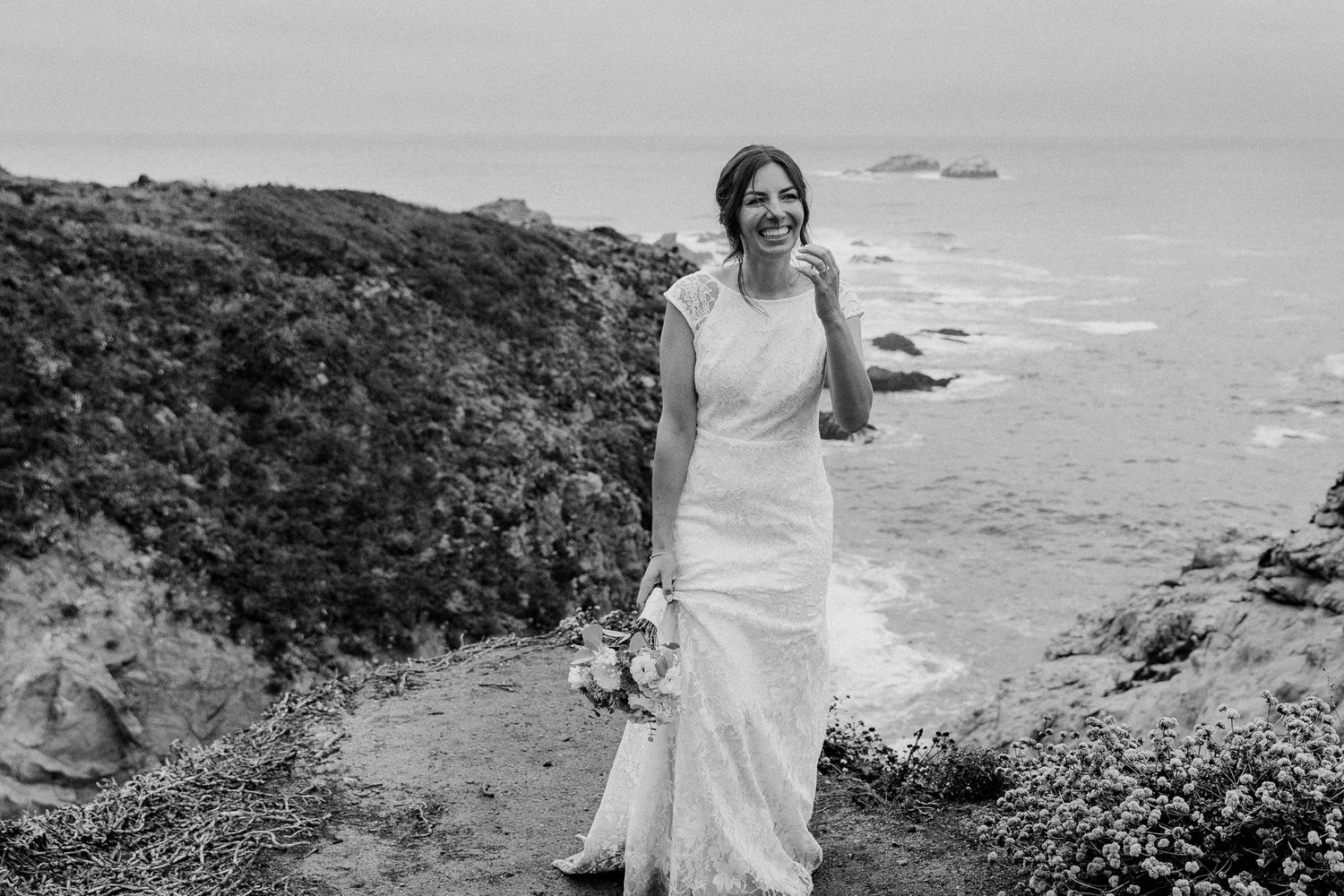 A bride in white dress holding a bouquet walks along a coastal path with ocean views.