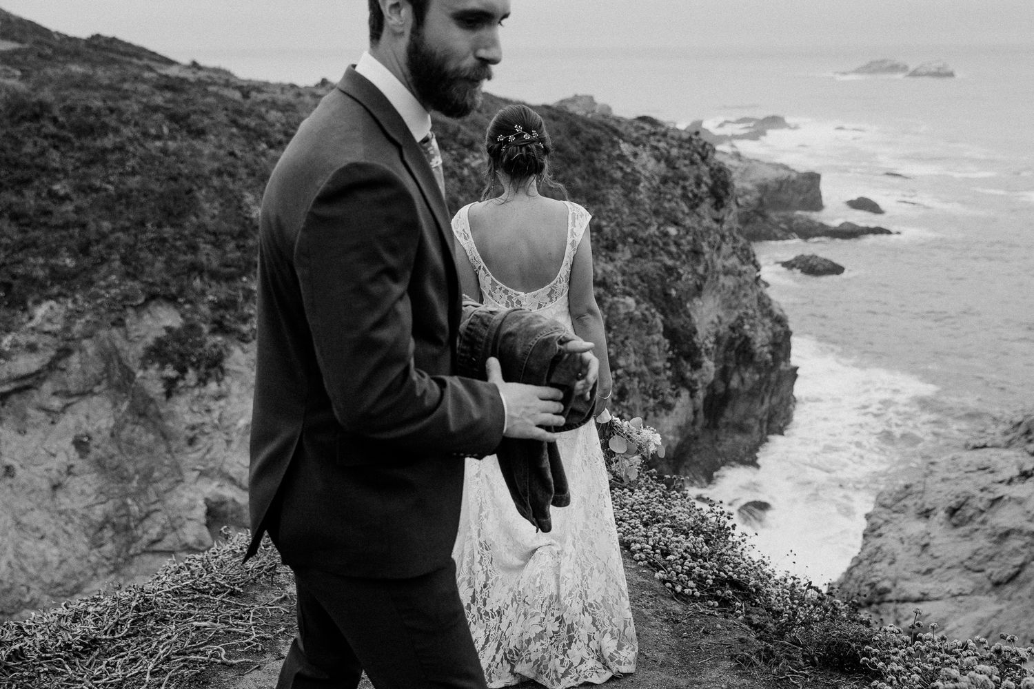 A wedding couple embraces while standing on rugged coastal cliffs during an overcast day.