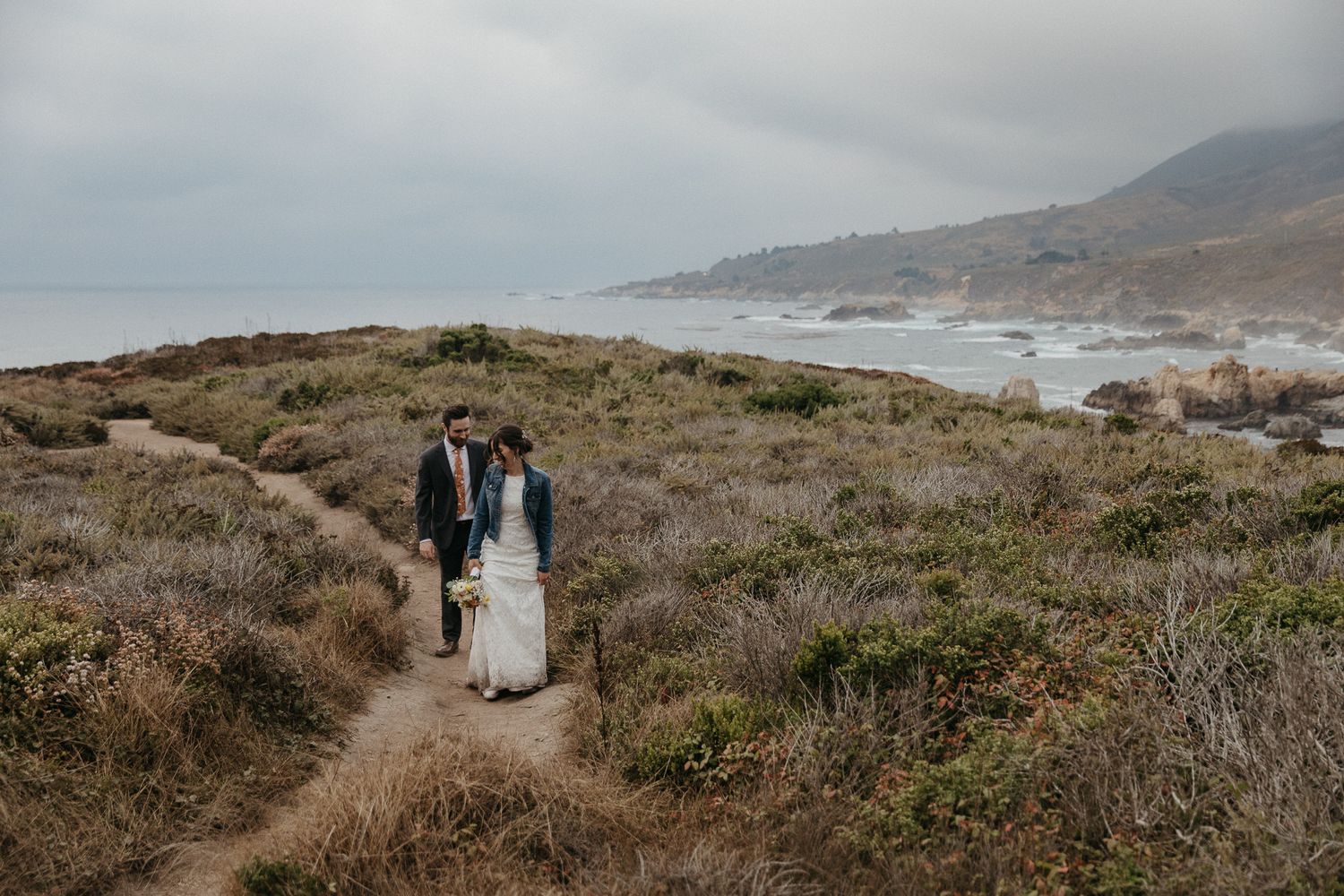 A couple walks along a winding coastal trail with dramatic ocean views in the background.