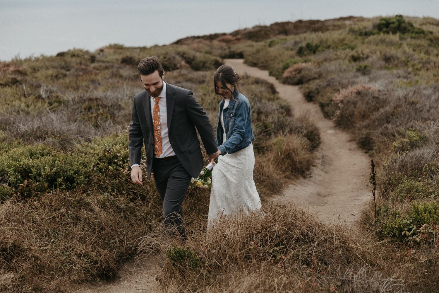 A couple explores a rustic dirt path through coastal brush on their wedding day.