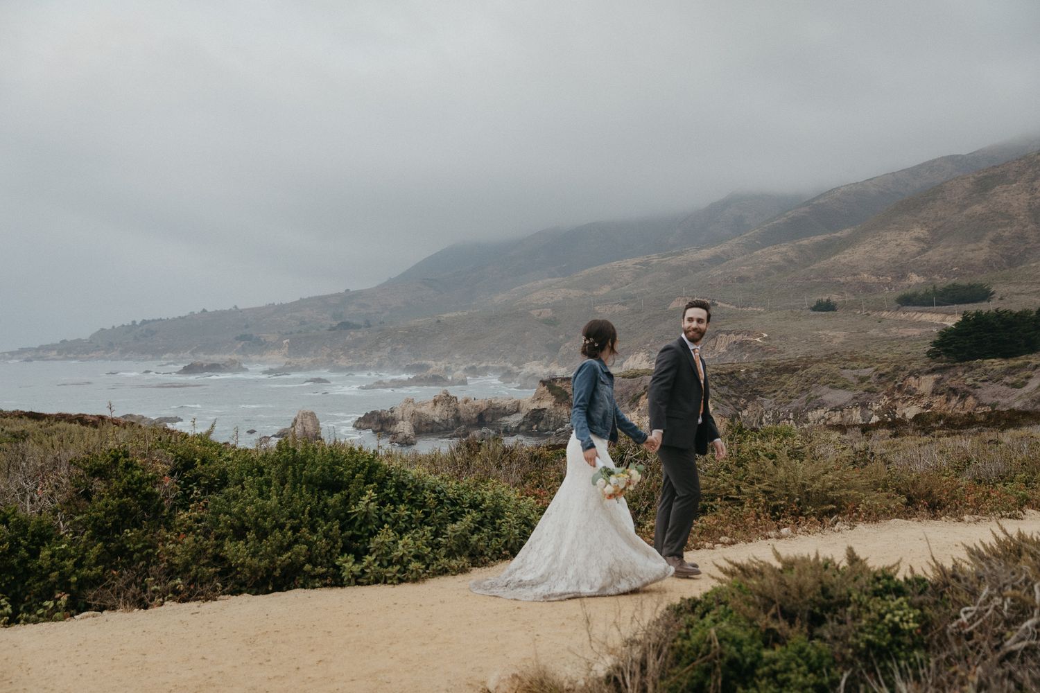 A couple poses on a scenic coastal path with fog-covered mountains and ocean in the background.