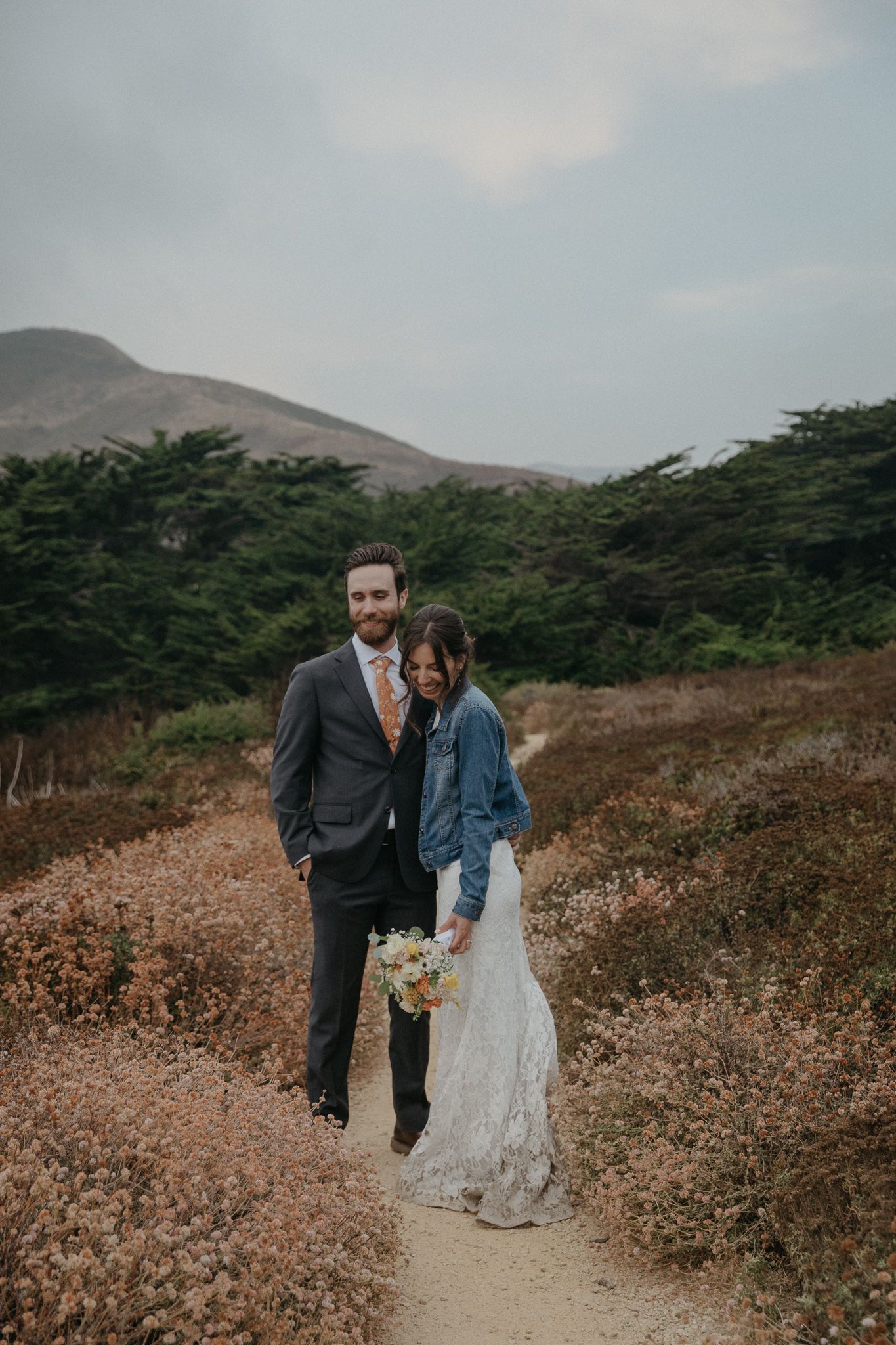 A couple shares a tender moment on a scenic coastal trail surrounded by cypress trees and wildflowers.