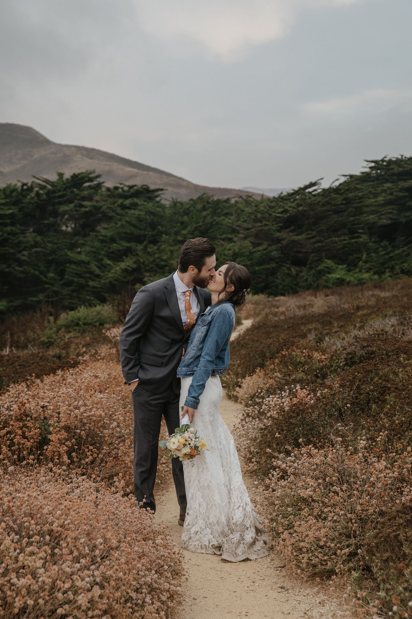 A romantic moment captured on a scenic coastal trail with mountains and cypress trees in the background.