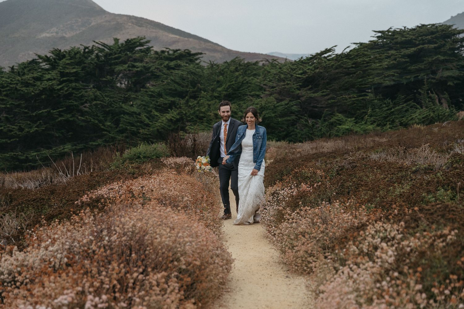 A couple walks hand in hand along a winding dirt path with mountains and cypress trees in the background.