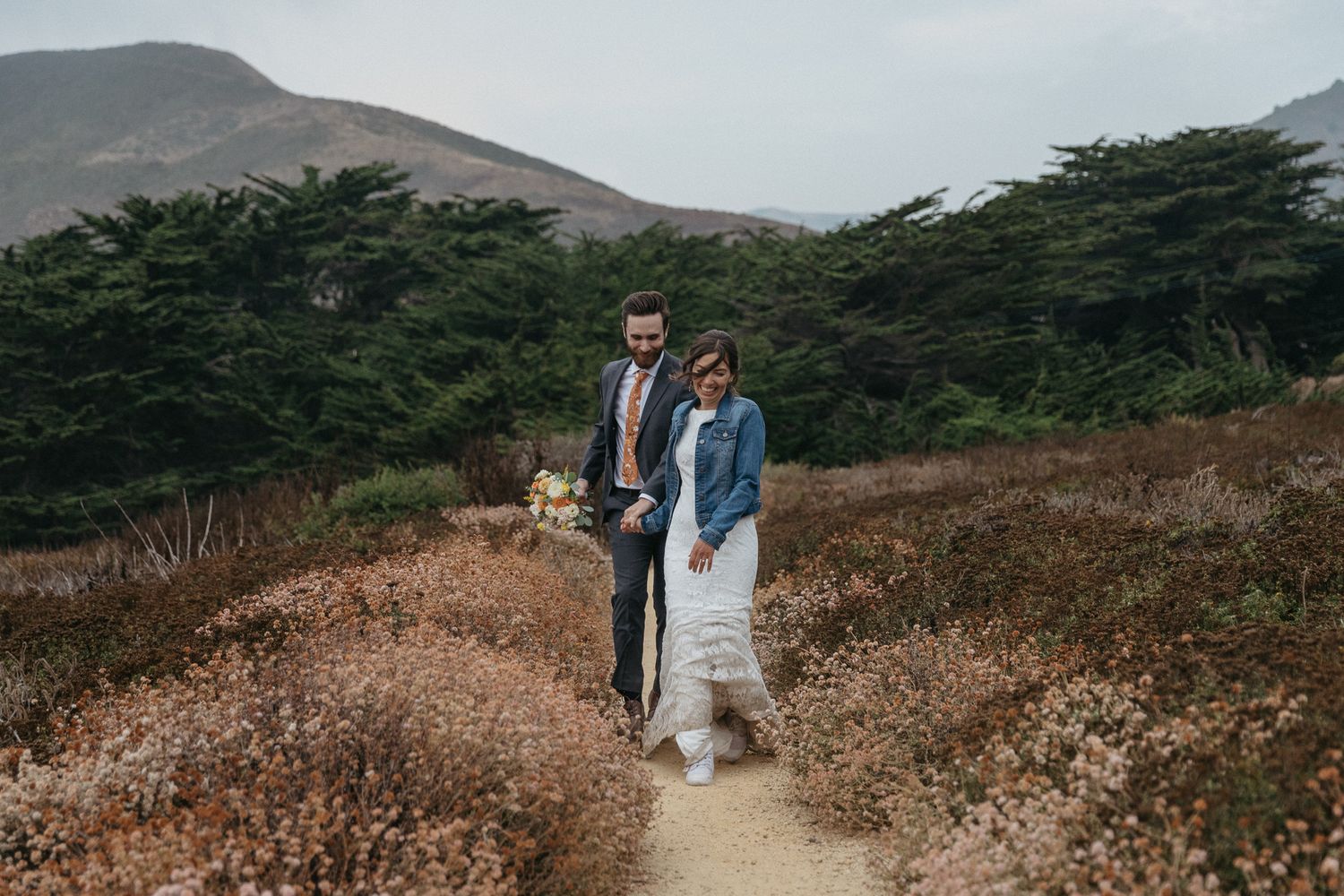 A couple strolls along a coastal path lined with dried wildflowers and cypress trees.