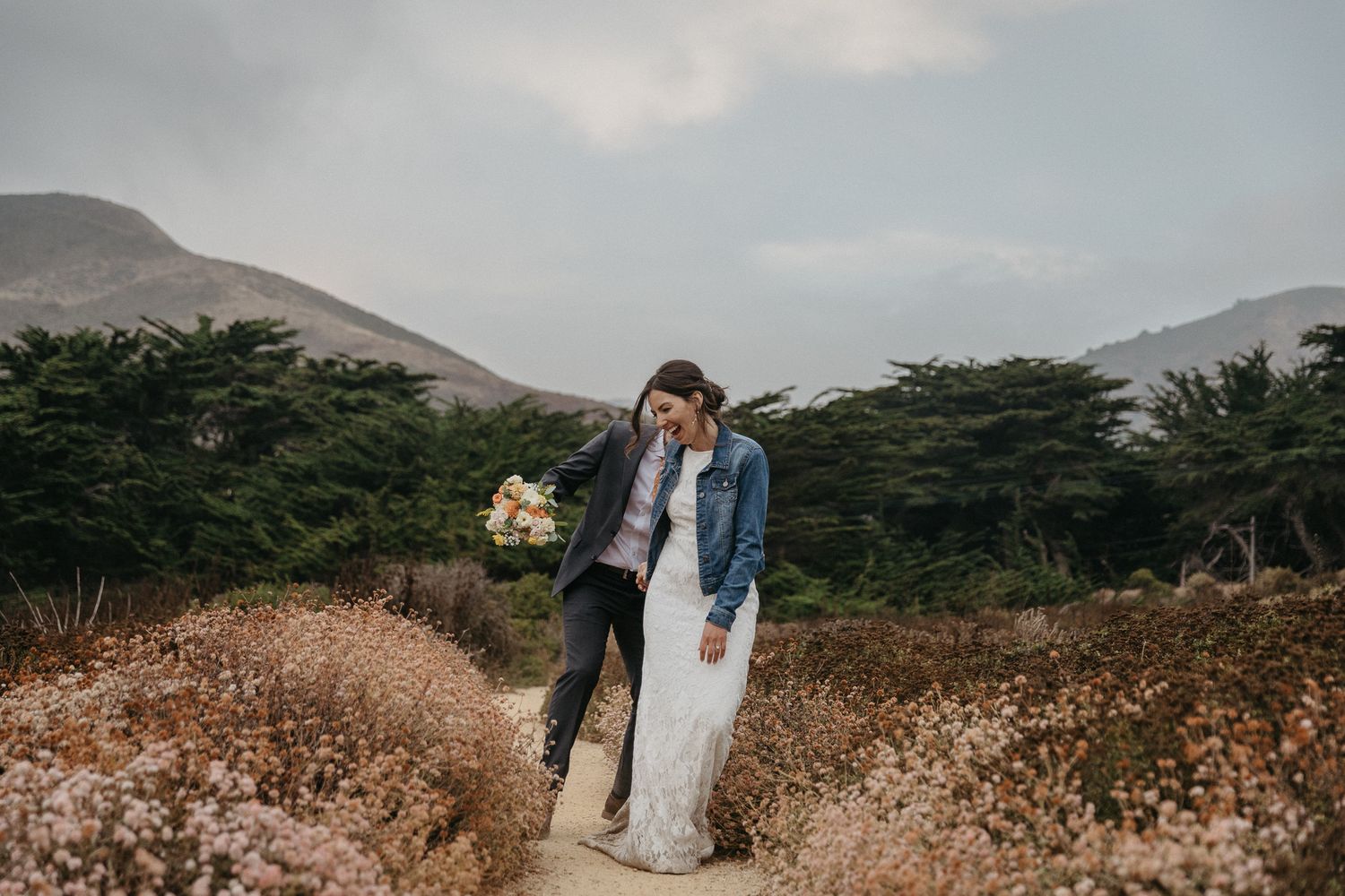 A couple embraces on a nature trail surrounded by coastal vegetation and mountains.