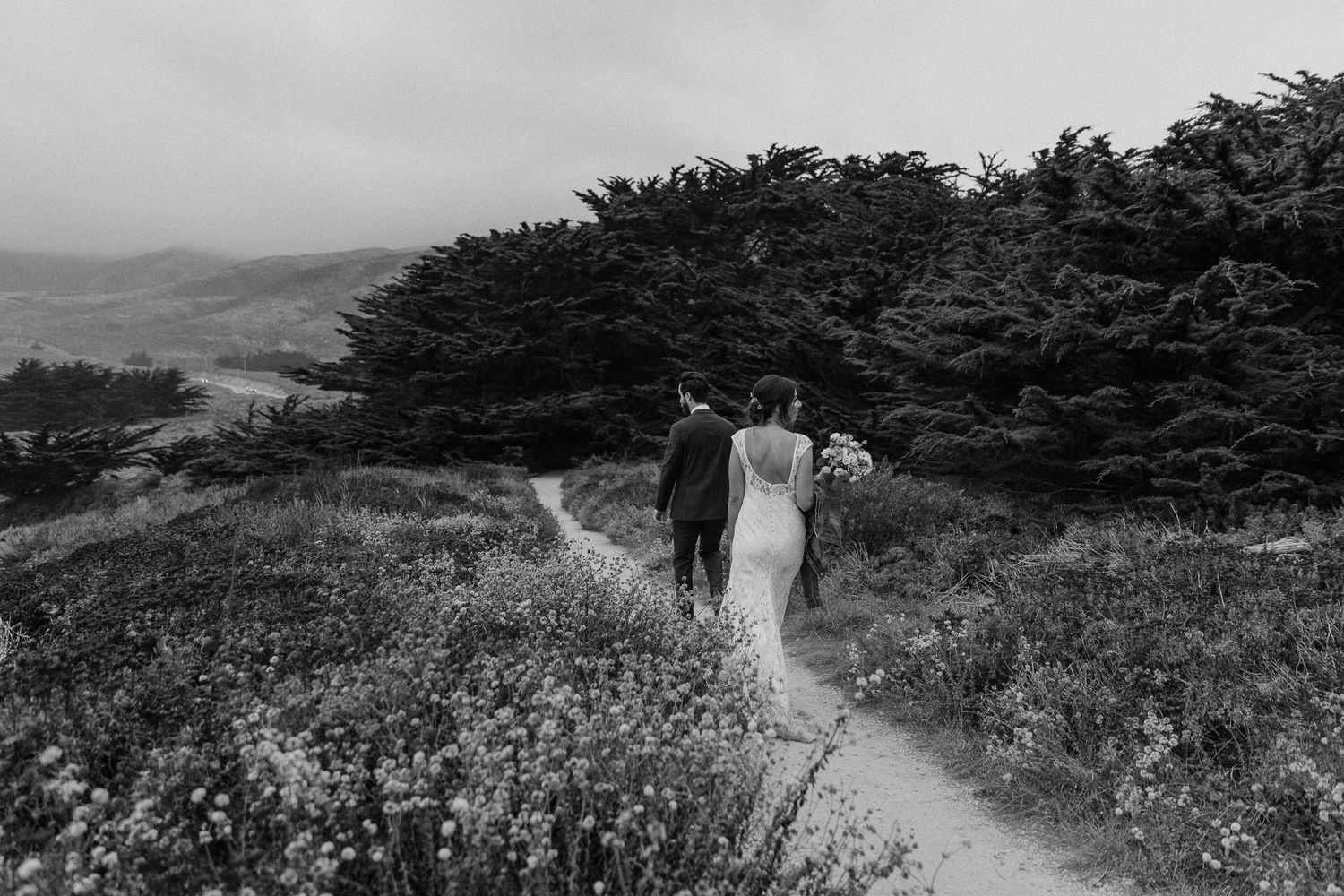 A black and white photo of a couple walking along a wildflower-lined coastal path.