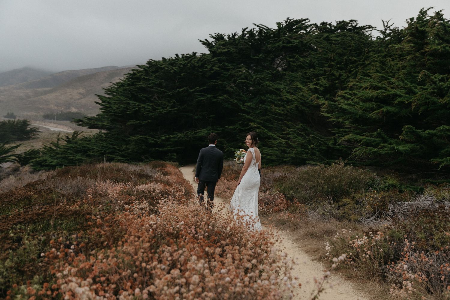 A couple walks down a misty coastal trail surrounded by wild coastal vegetation.