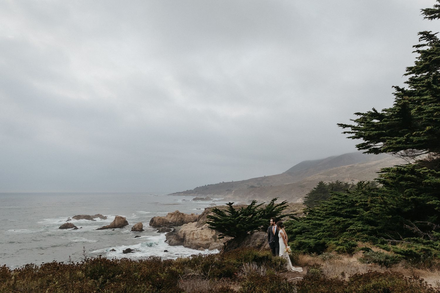 Dramatic coastal landscape with couple overlooking rocky Pacific shoreline and cypress trees.