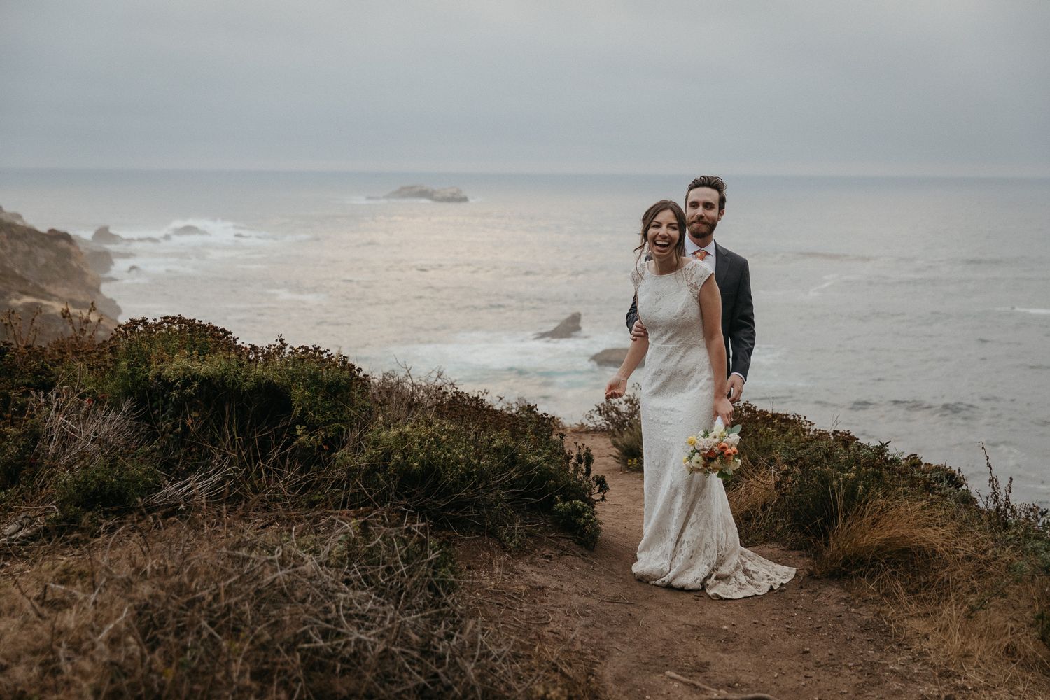 A couple stands on a cliff edge overlooking the dramatic Pacific coastline.