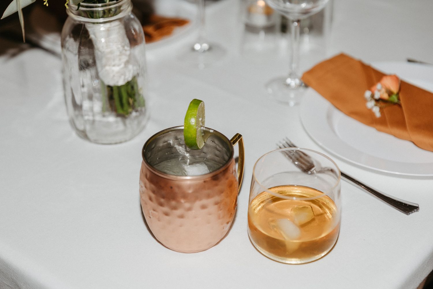 A copper Moscow mule mug and glass of whiskey on a white tablecloth with table settings.