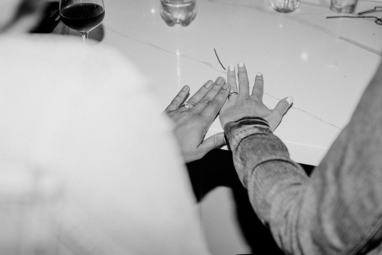 Black and white image of hands resting on a table near wine glasses.