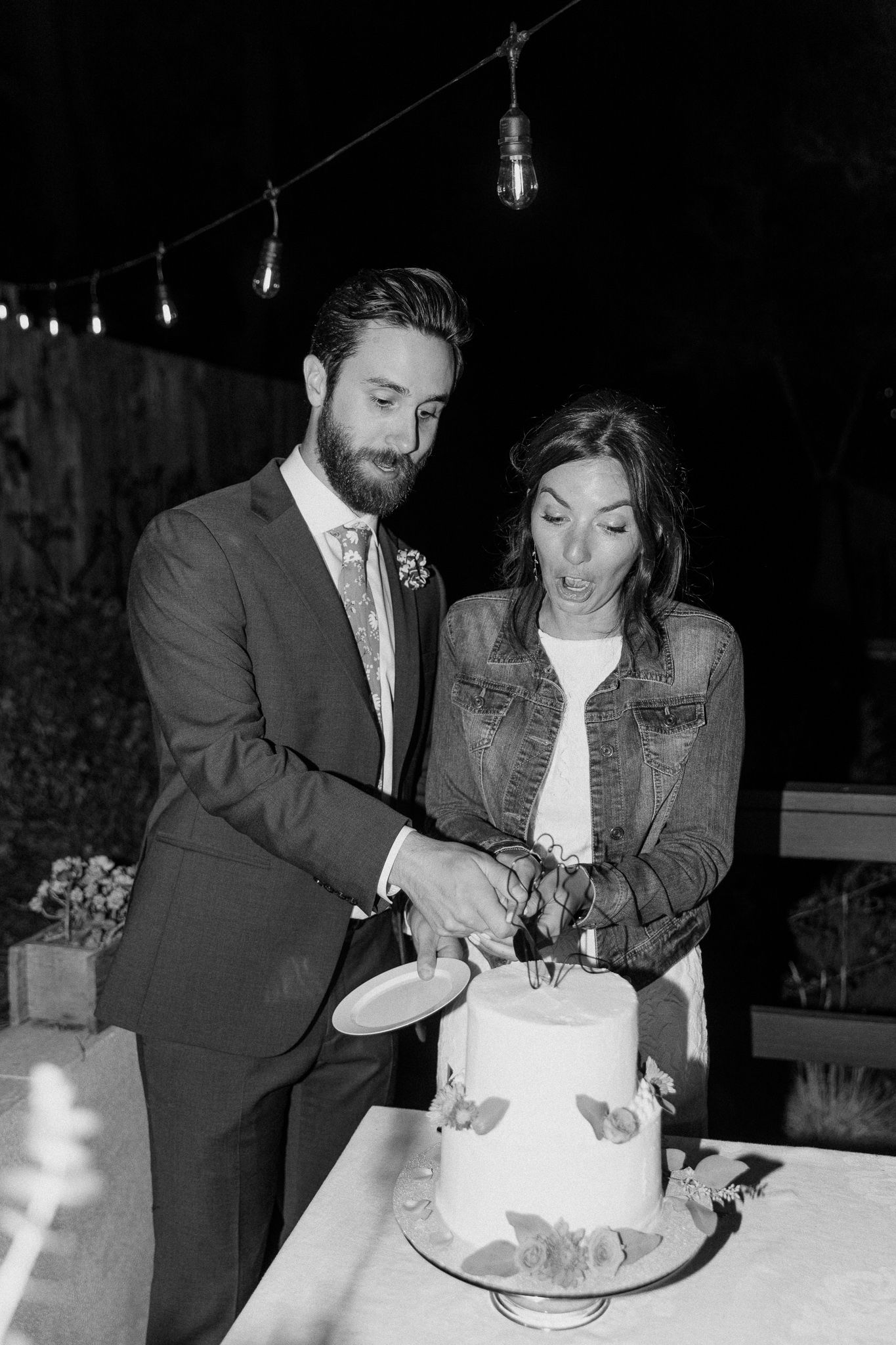 A couple in wedding attire cutting their white wedding cake together at an outdoor evening reception.
